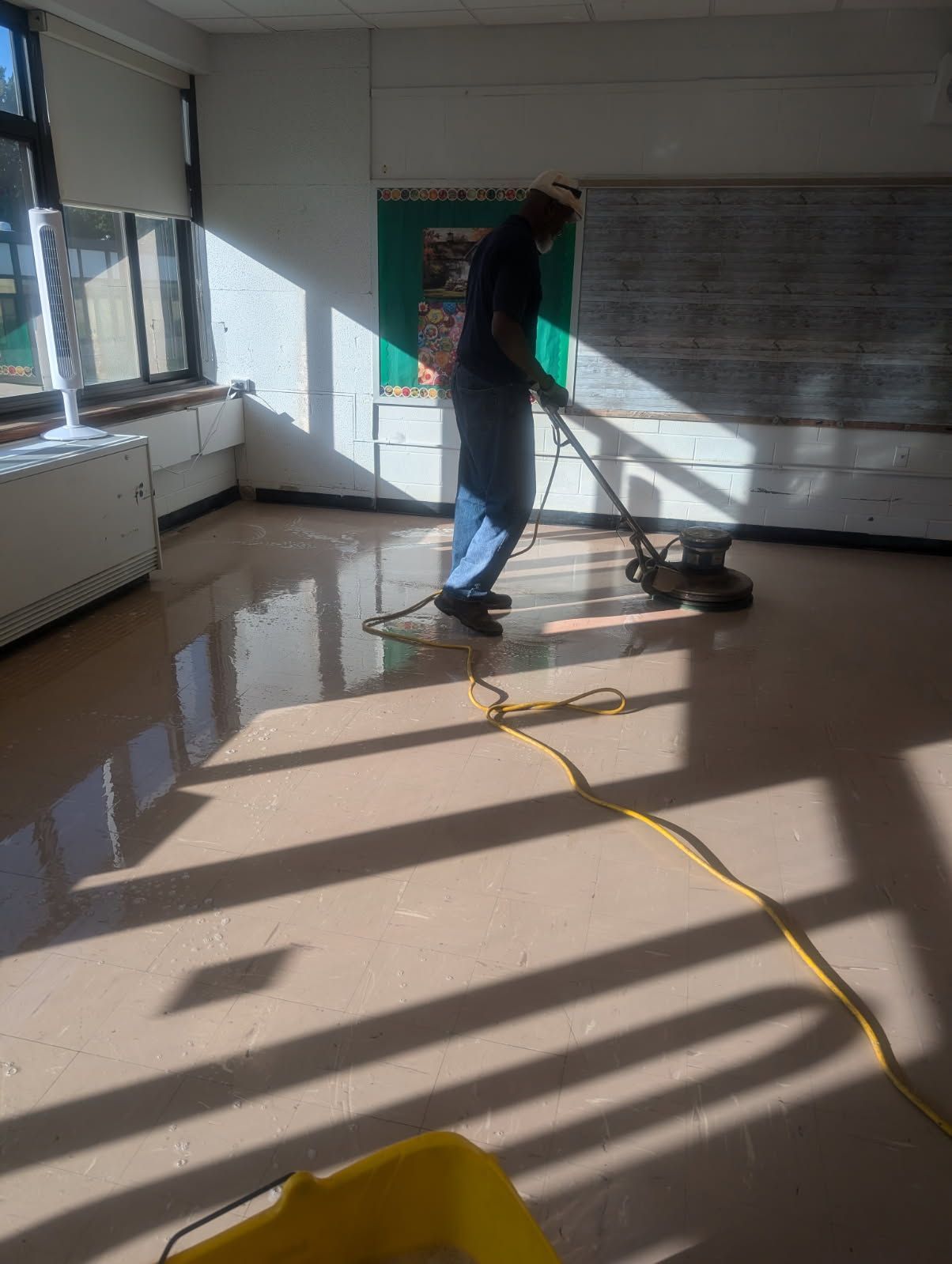 Person in a sunlit empty room cleaning a wet floor with a floor buffer, yellow cord across the floor.