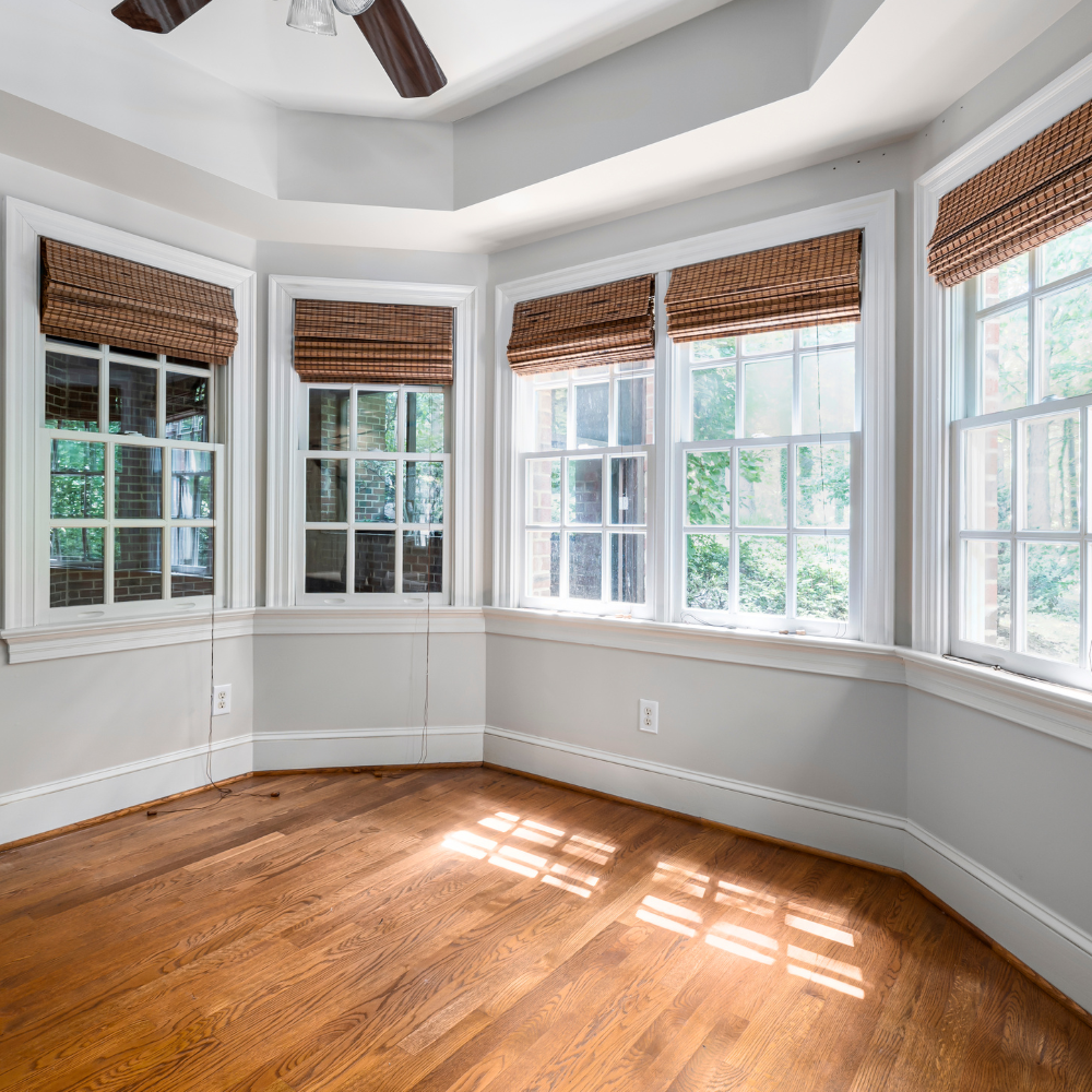 Empty, sunlit room with hardwood floors, white walls, and bay windows with brown shades.