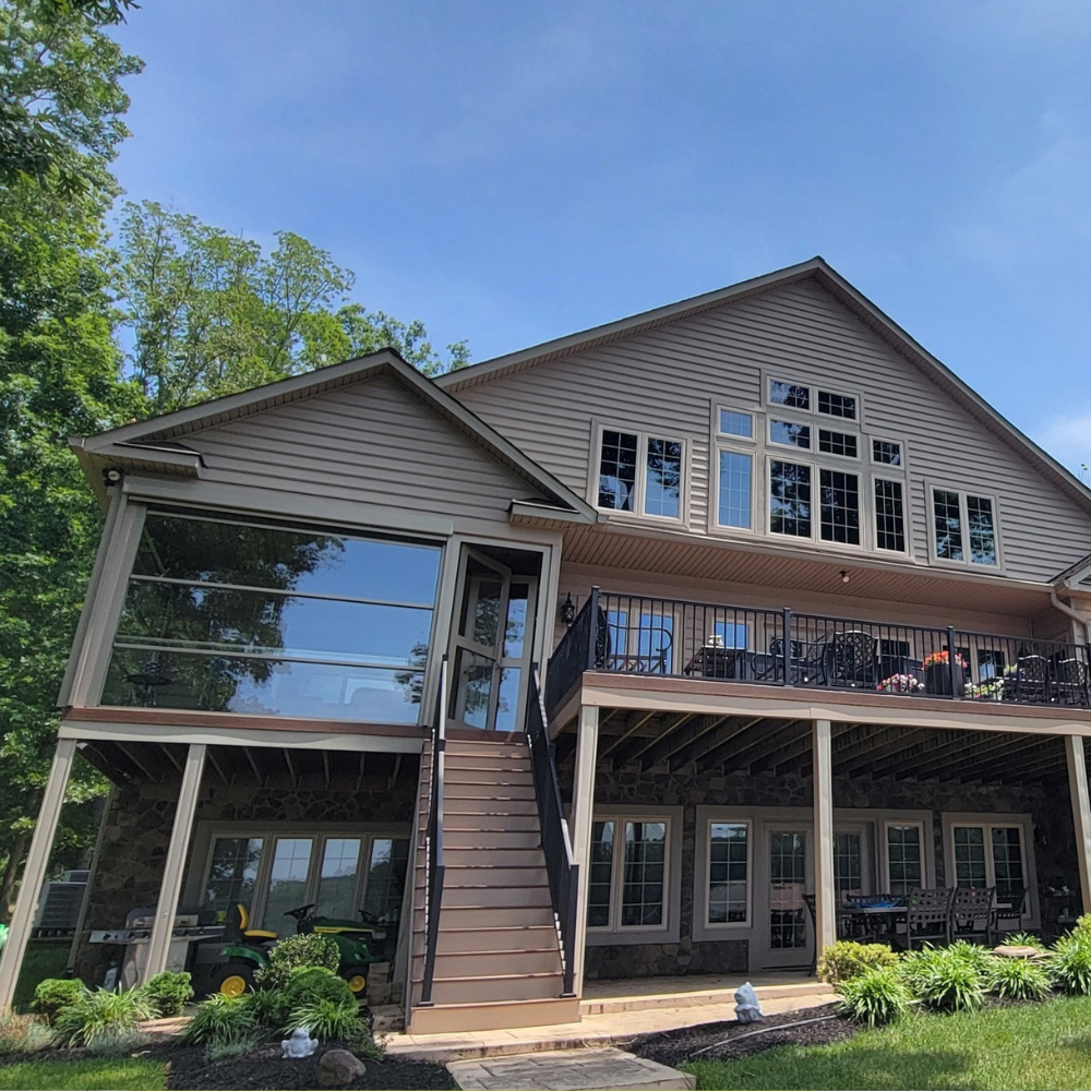 Two-story tan house with large windows, balconies, and a glass-walled side addition, set against a blue sky.