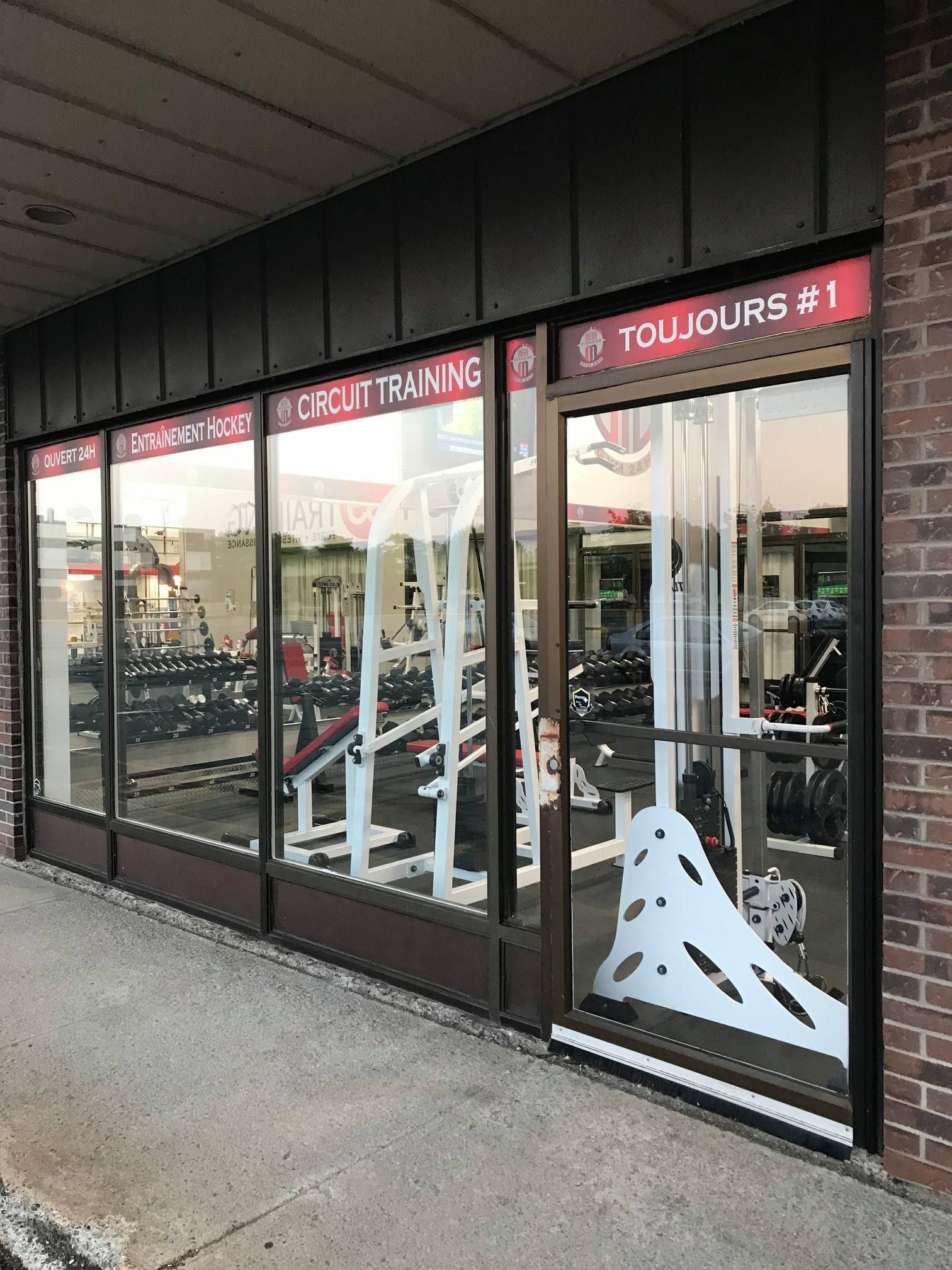 Façade de salle de sport avec de grandes vitrines, des appareils de musculation blancs à l'intérieur et une enseigne rouge indiquant « TOLLOXUS #1 »