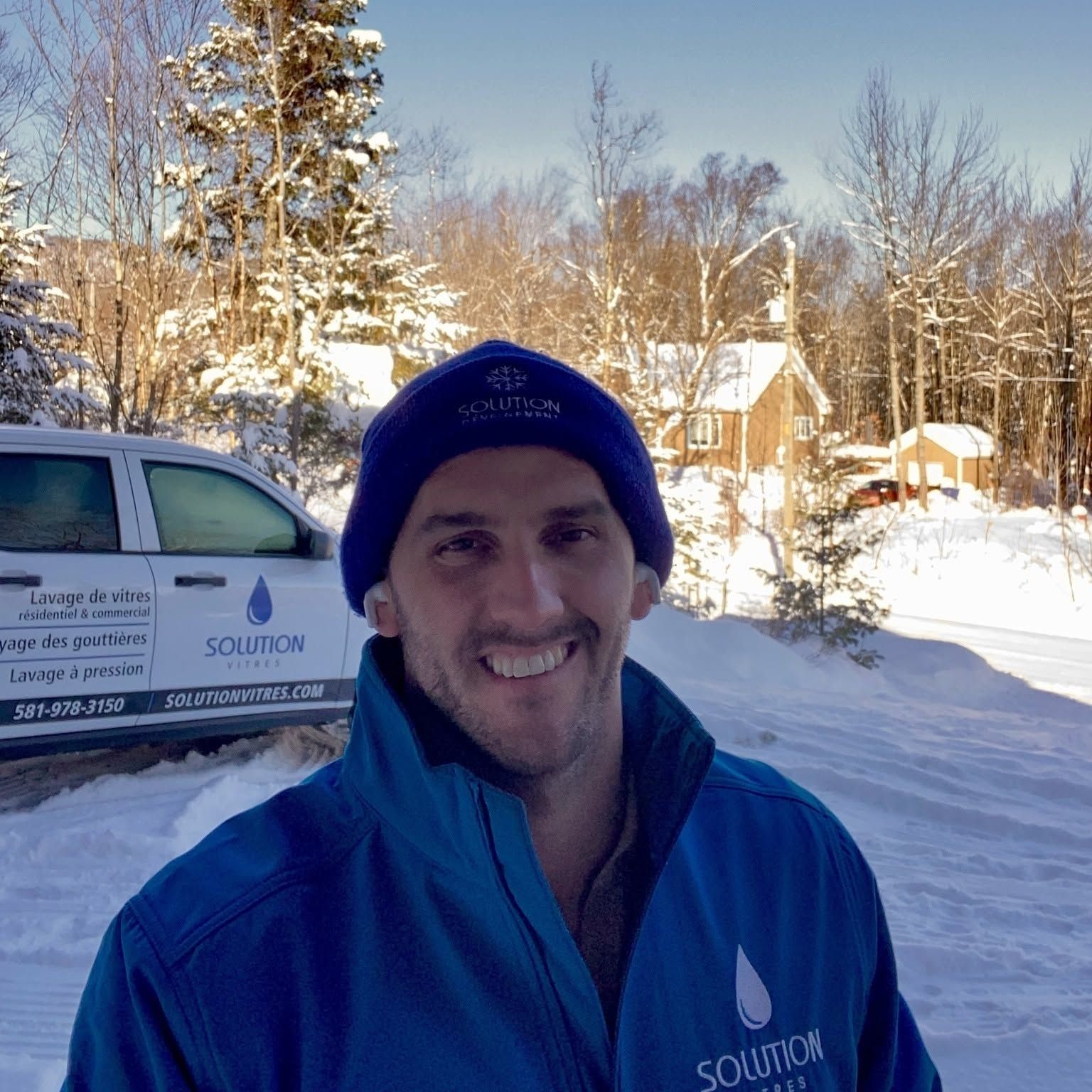 Un homme souriant, vêtu d'une veste bleue et d'un bonnet, se tient debout dans un quartier enneigé, à côté d'un camion de chantier.
