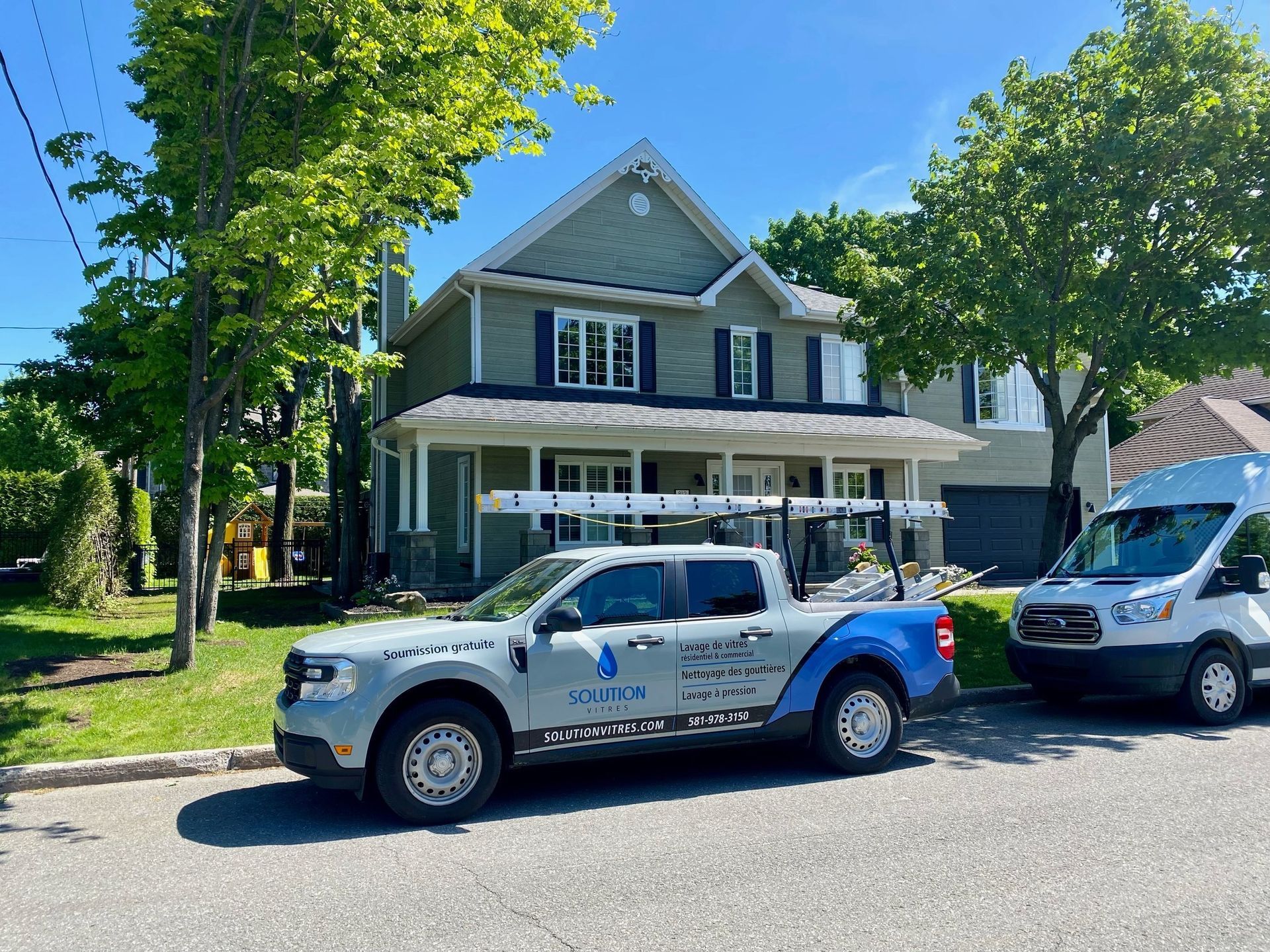 Camionnette blanche garée devant une maison grise dans une rue ensoleillée bordée d'arbres
