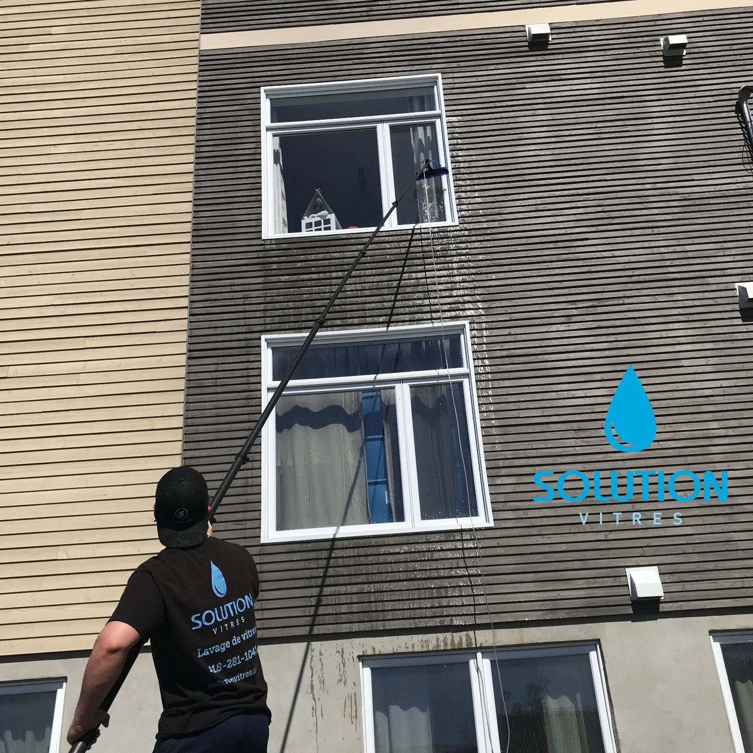Un ouvrier nettoie la façade d'un bâtiment au nettoyeur haute pression à l'aide d'une longue perche ; on aperçoit les fenêtres et le logo bleu d'une entreprise de nettoyage.