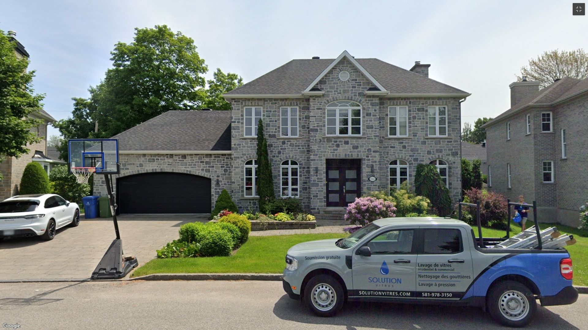 Maison de banlieue en pierre avec deux véhicules garés devant et une pelouse près de l'allée.