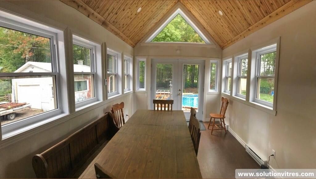 Véranda lumineuse avec une longue table, de grandes fenêtres et un plafond voûté en bois.