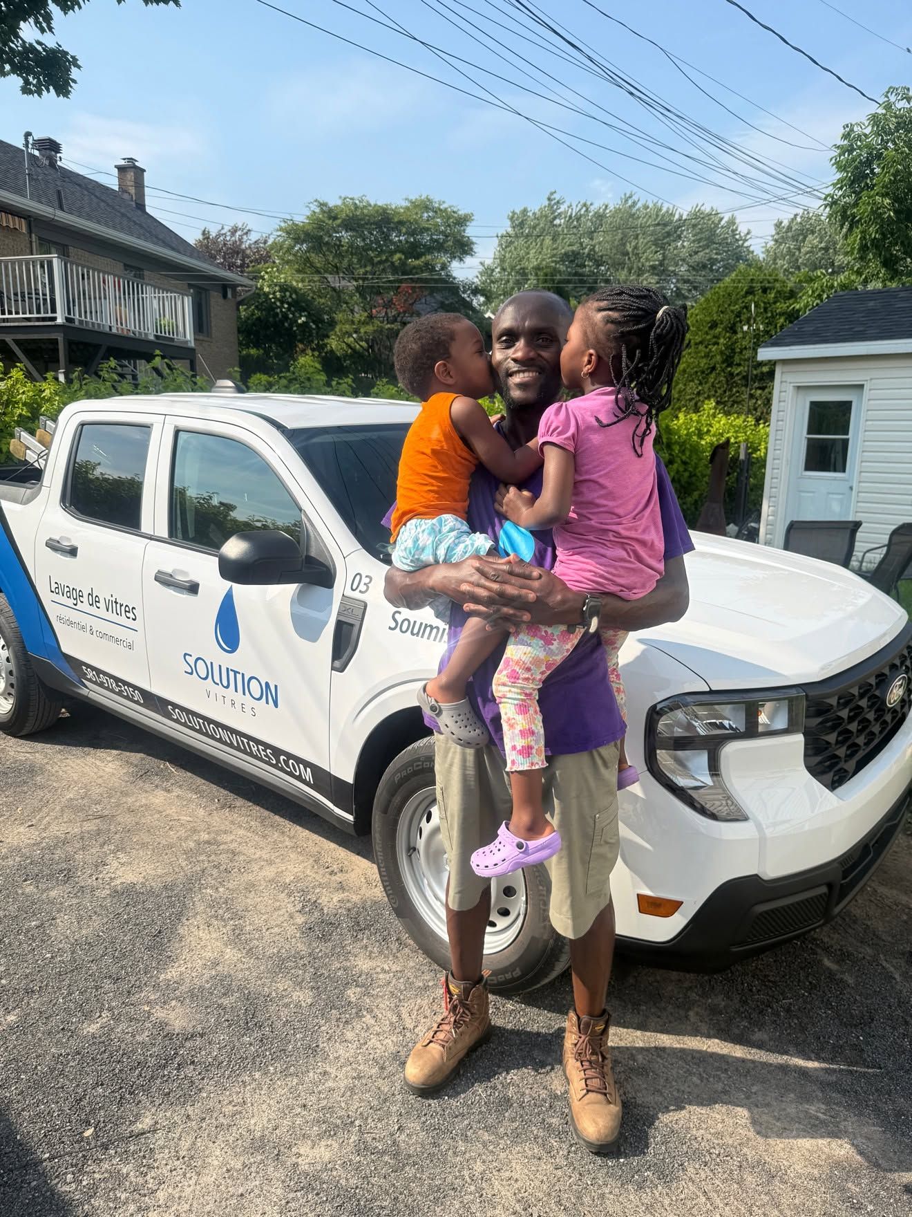 Un homme tenant deux enfants devant une camionnette blanche dans une rue ensoleillée