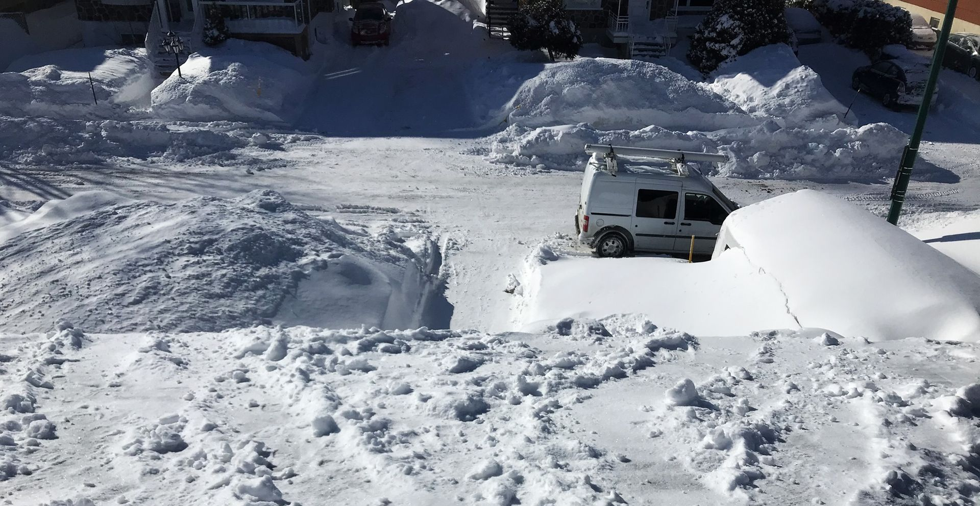 Rue enneigée avec une camionnette blanche partiellement ensevelie sous une épaisse couche de neige après de fortes chutes de neige.