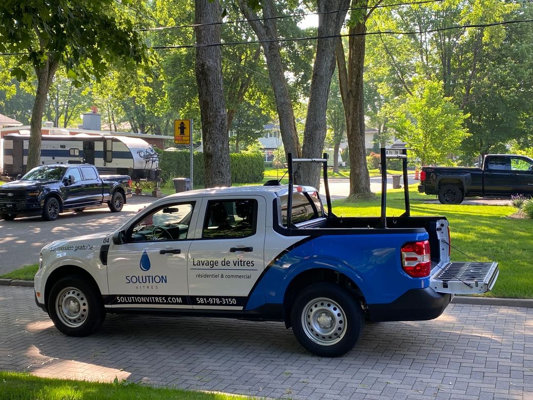 Une camionnette blanche et bleue avec des barres de toit est garée dans une rue bordée d'arbres.