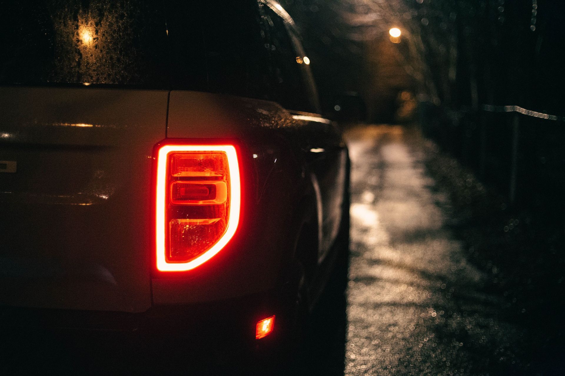 Close-up of a dark car’s red taillight glowing on a wet road at night