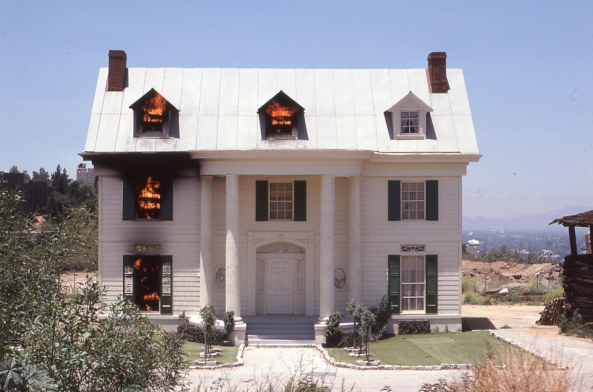 White two-story house with lit dormer windows, black shutters, and a half the room on fire.