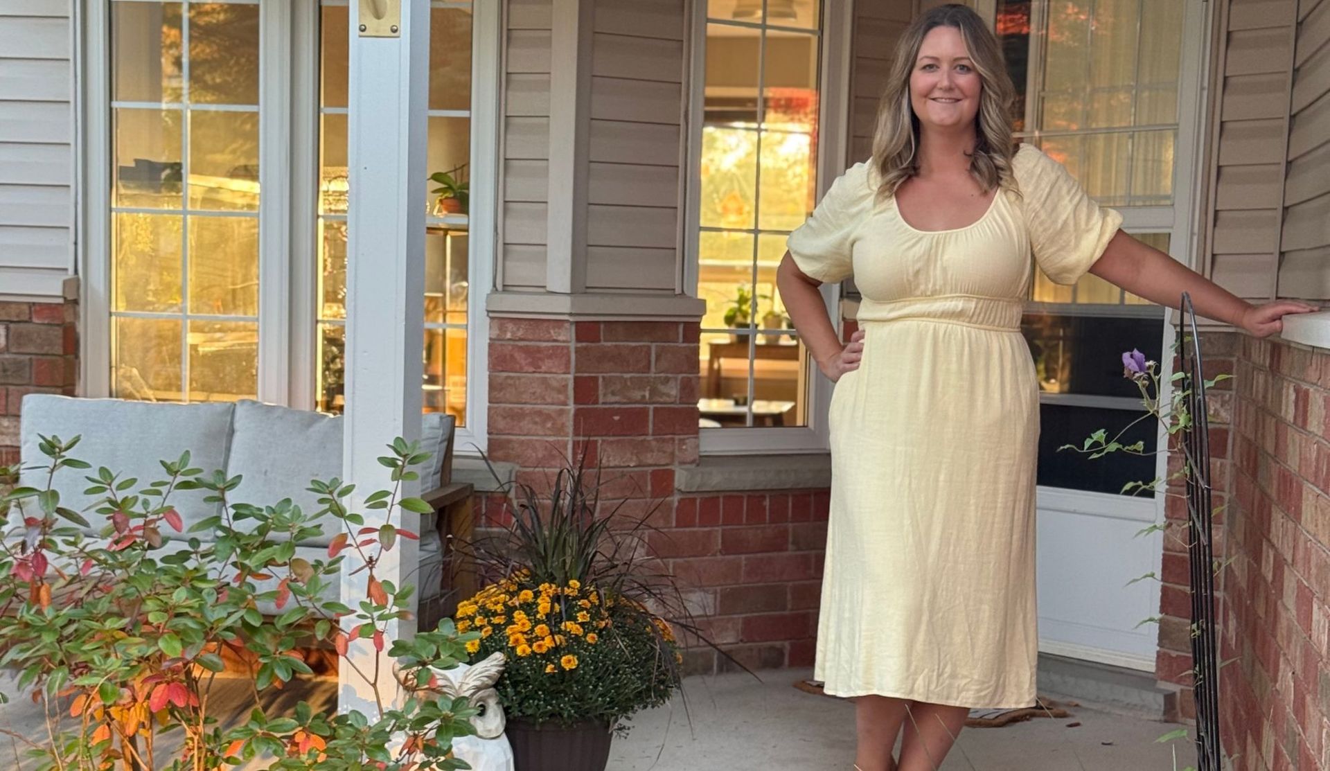 Woman in a pale yellow dress standing on a porch beside potted flowers, smiling at the camera.