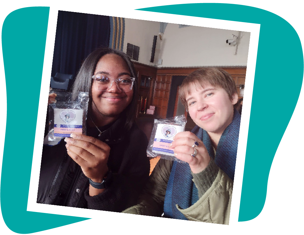 Two smiling women holding up clear packets with pink labels. Inside, a room with dark wood paneling is visible.