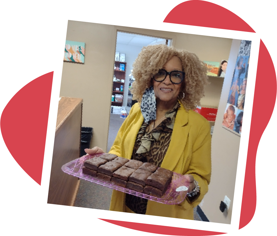Woman in yellow jacket holds a tray of brownies, smiling in an office setting.