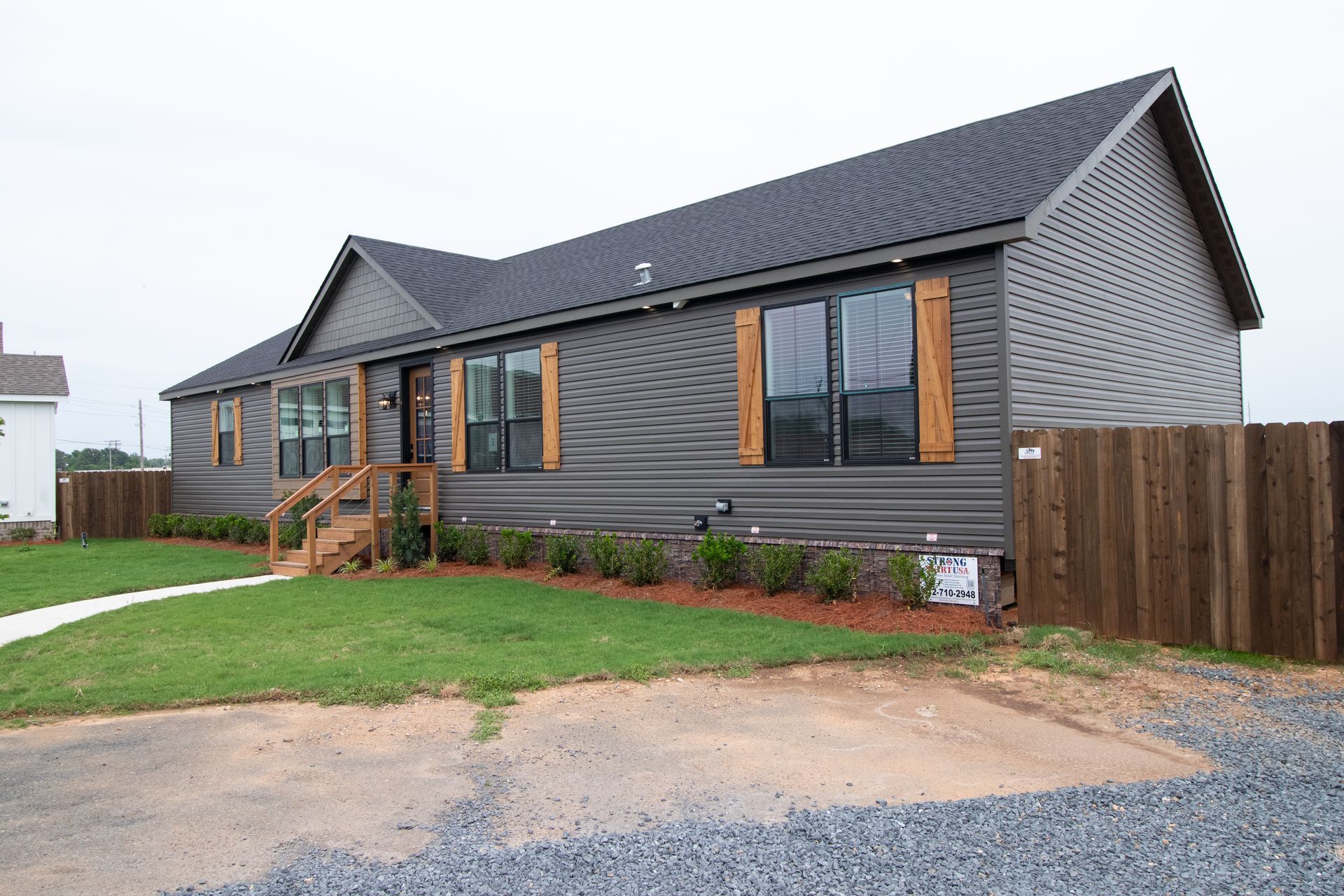 Modern gray single-story house with dark roof, wood steps, and a small front lawn by a gravel drive