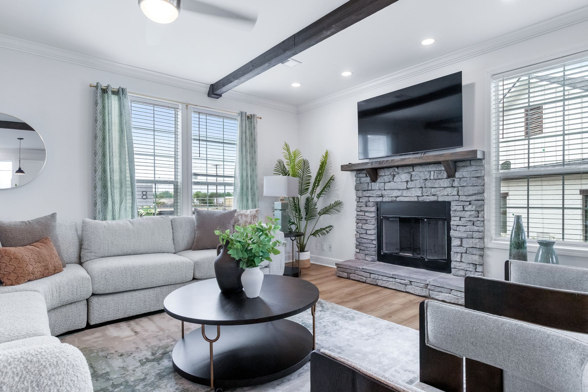 Bright modern living room with gray sectional, stone fireplace, large windows, and a wall-mounted TV