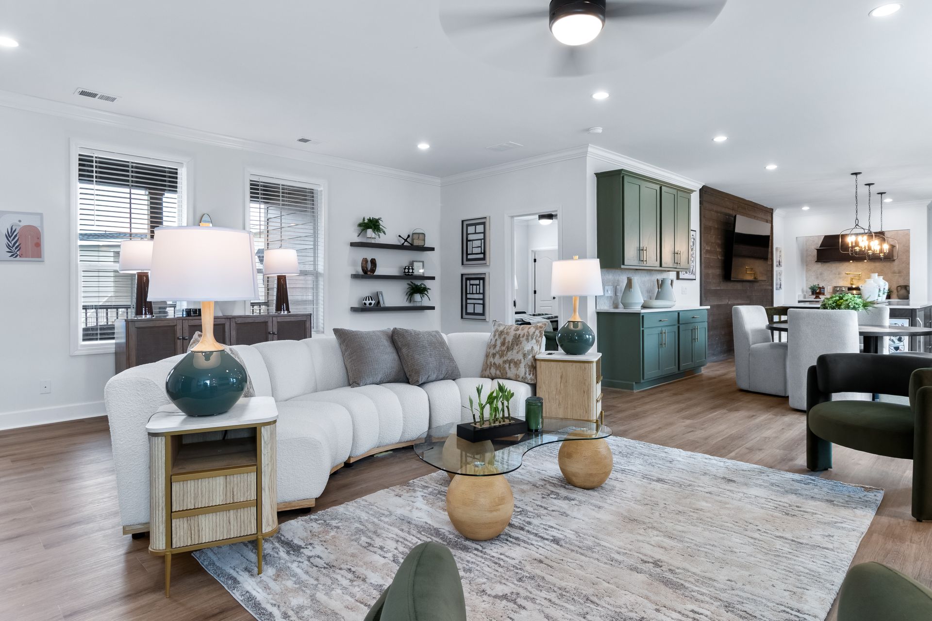 Bright open-concept living room with white sofa, wood floors, teal accents, and kitchen in background