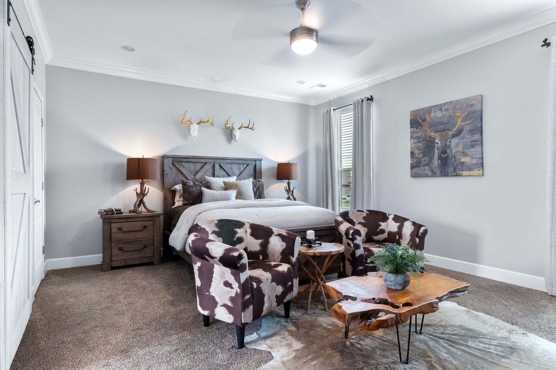 Bright bedroom with a bed, two patterned chairs, wood nightstands, and a glass coffee table on a gray carpet.