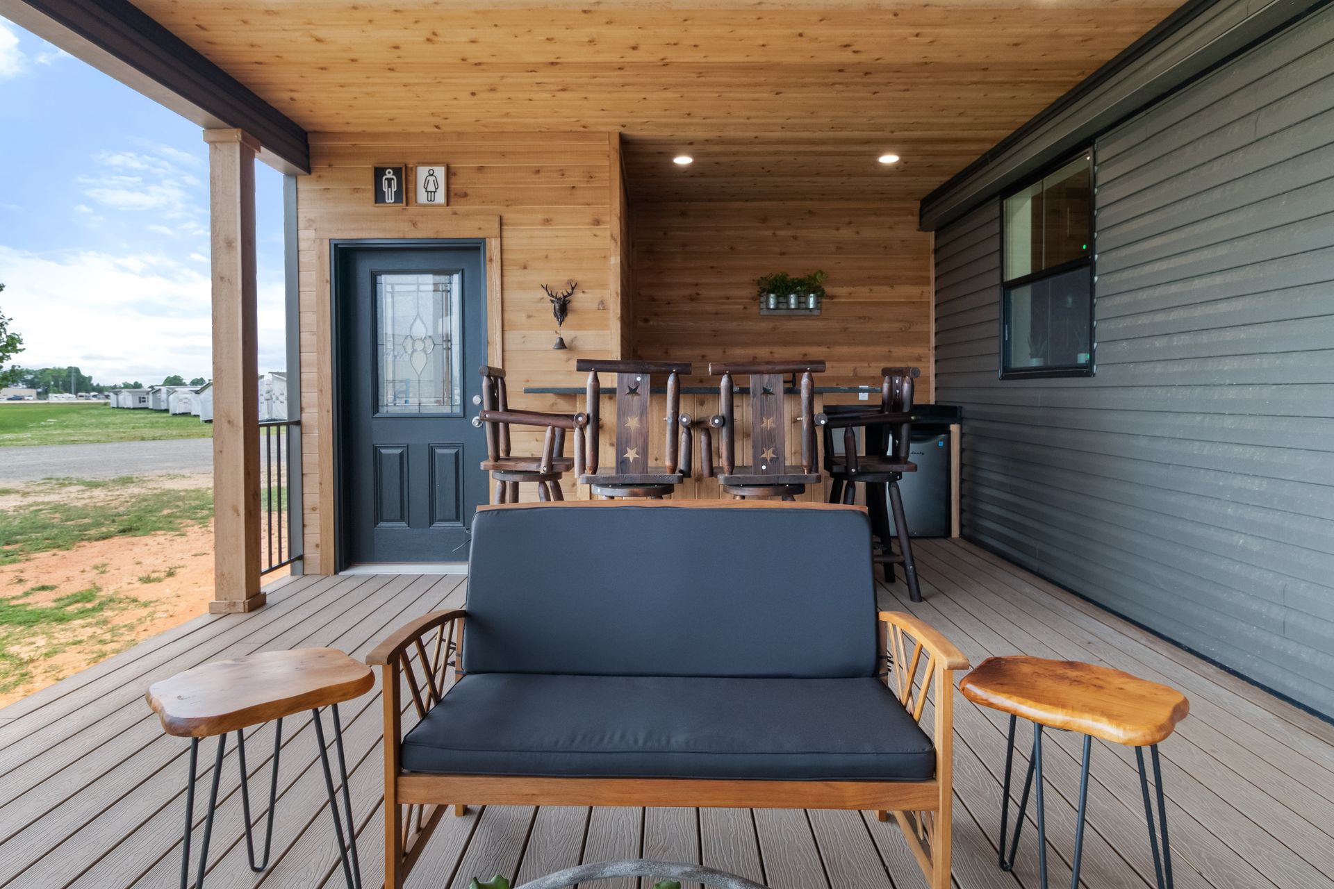 Covered porch with black sofa, wooden chairs, and a gray door beside a grassy yard.
