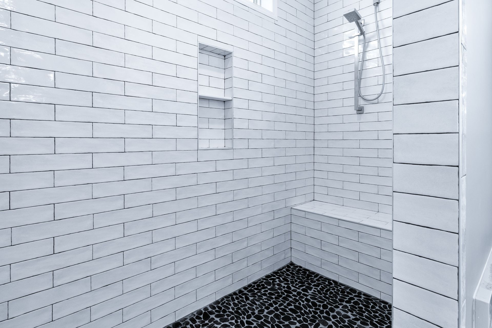 Empty white-tiled shower with built-in shelf, handheld showerhead, and black pebble floor.