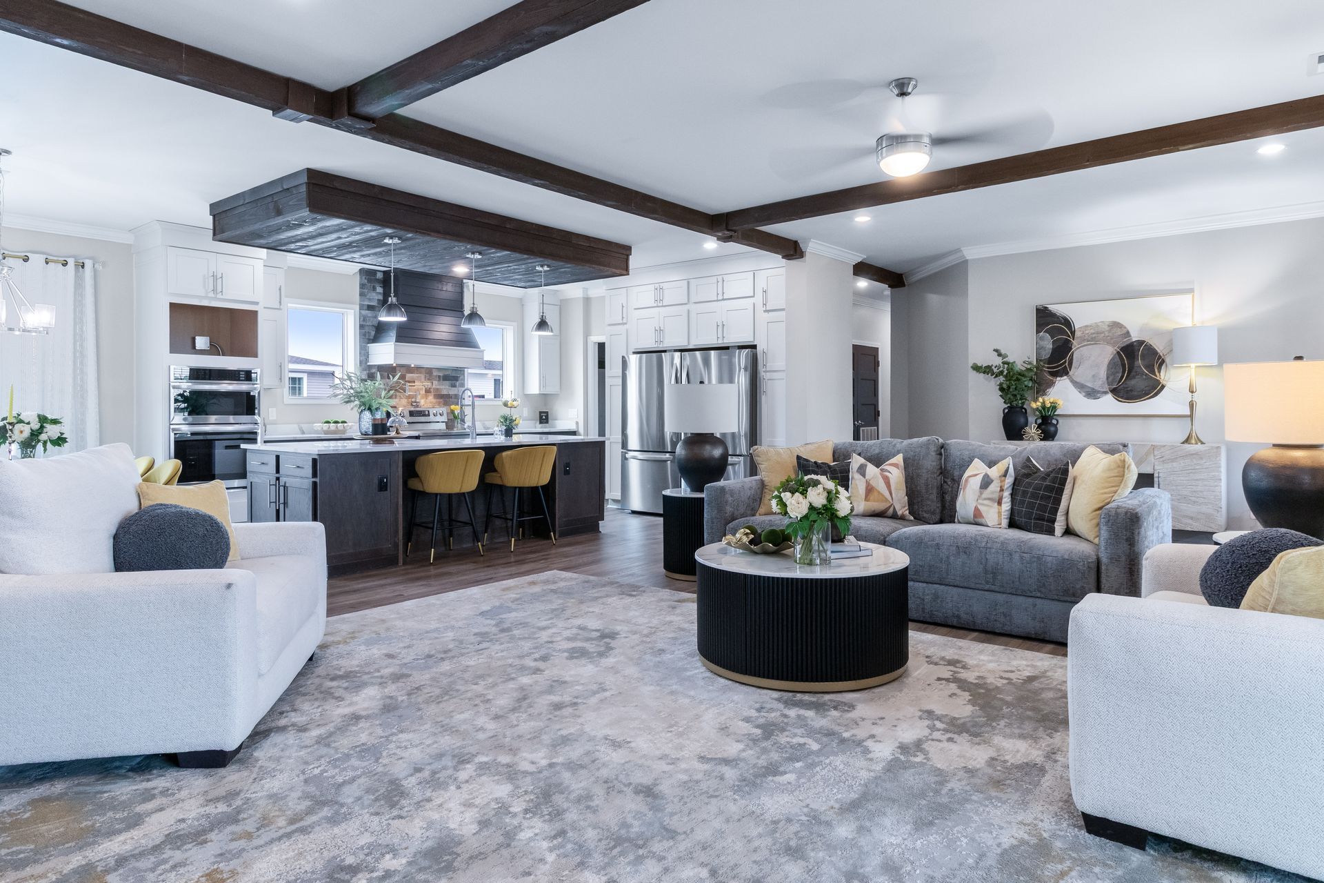Modern open-concept living room with gray sofas, round black coffee table, kitchen, and exposed ceiling beams