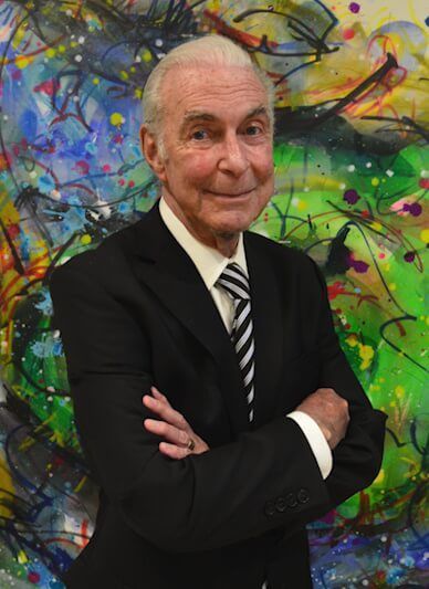 An older man in a suit and tie stands with arms crossed against a colorful, abstract painting background.