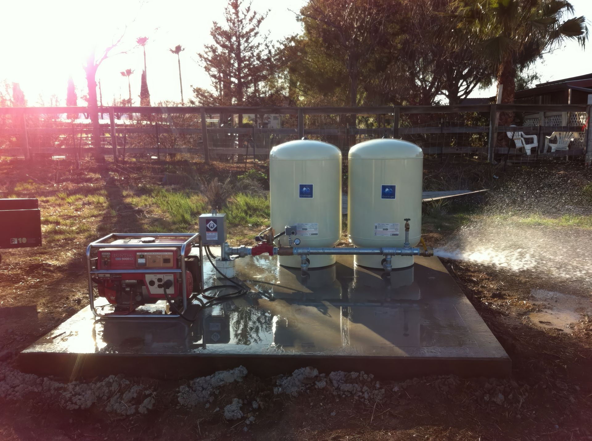 Two white tanks on a platform with a pump spraying water outdoors at sunset