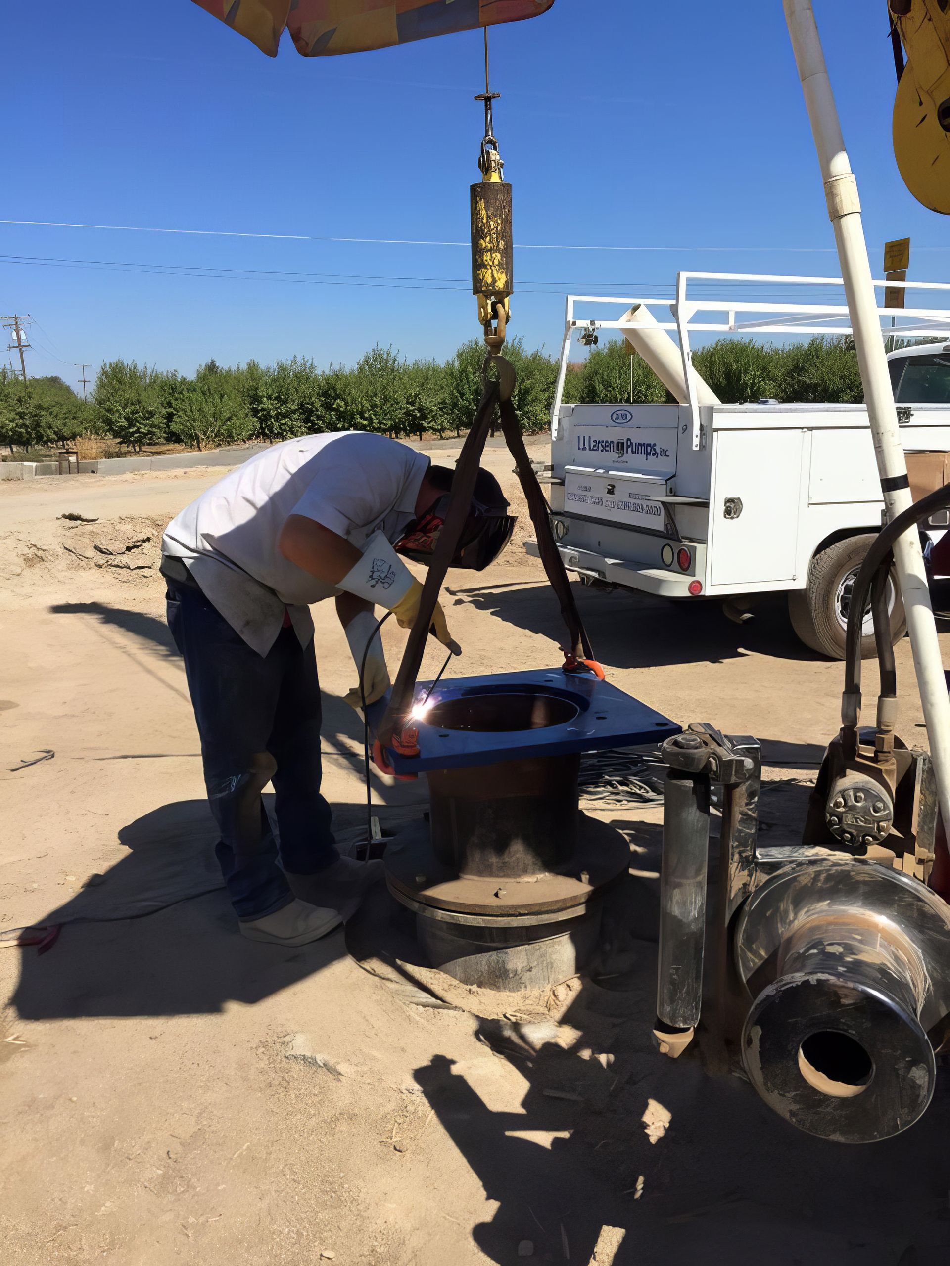 Worker bending over a blue well rig in a sandy outdoor worksite beside a white truck