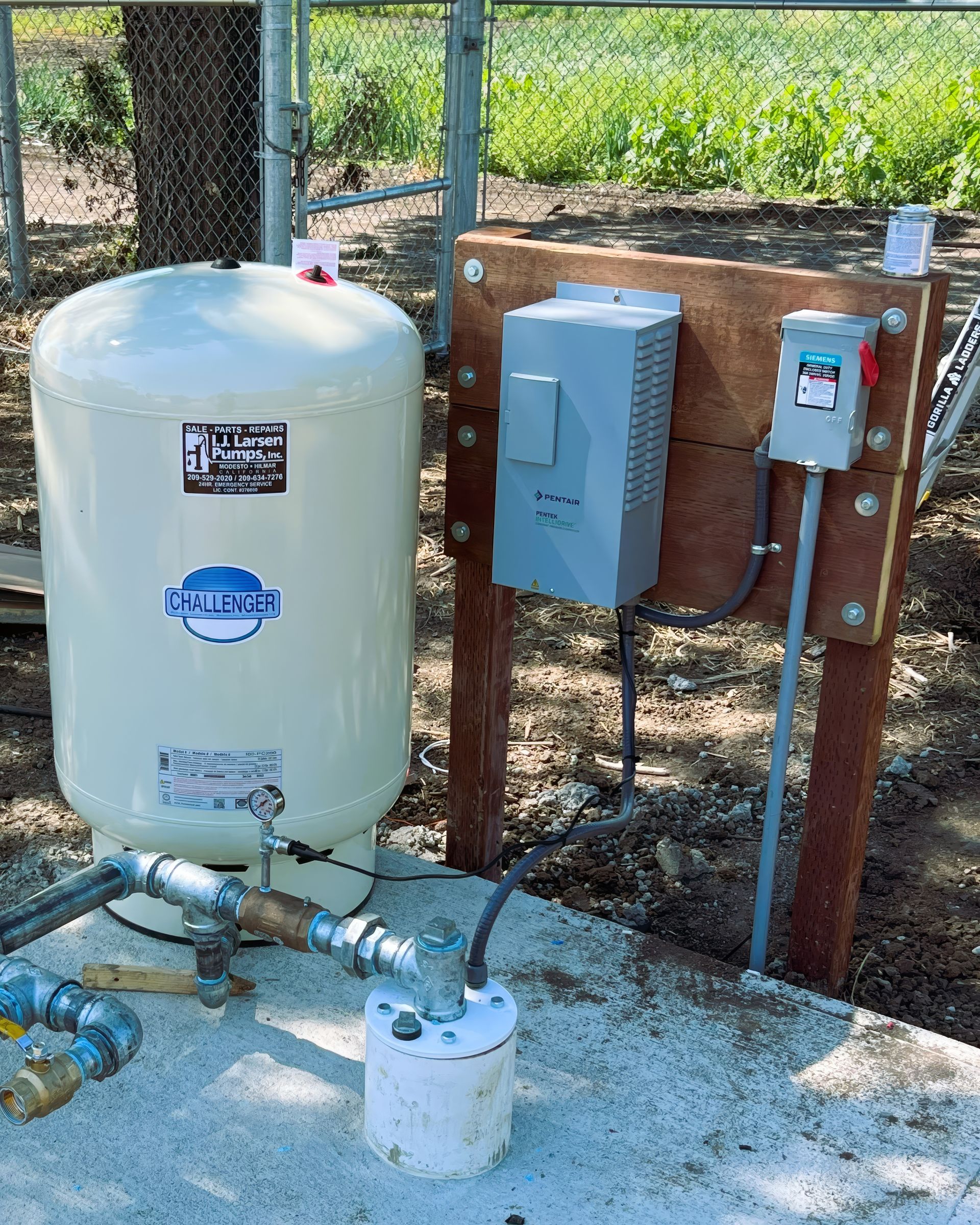 Outdoor water tank with pipes, gauges, and electrical boxes mounted on a wooden frame.