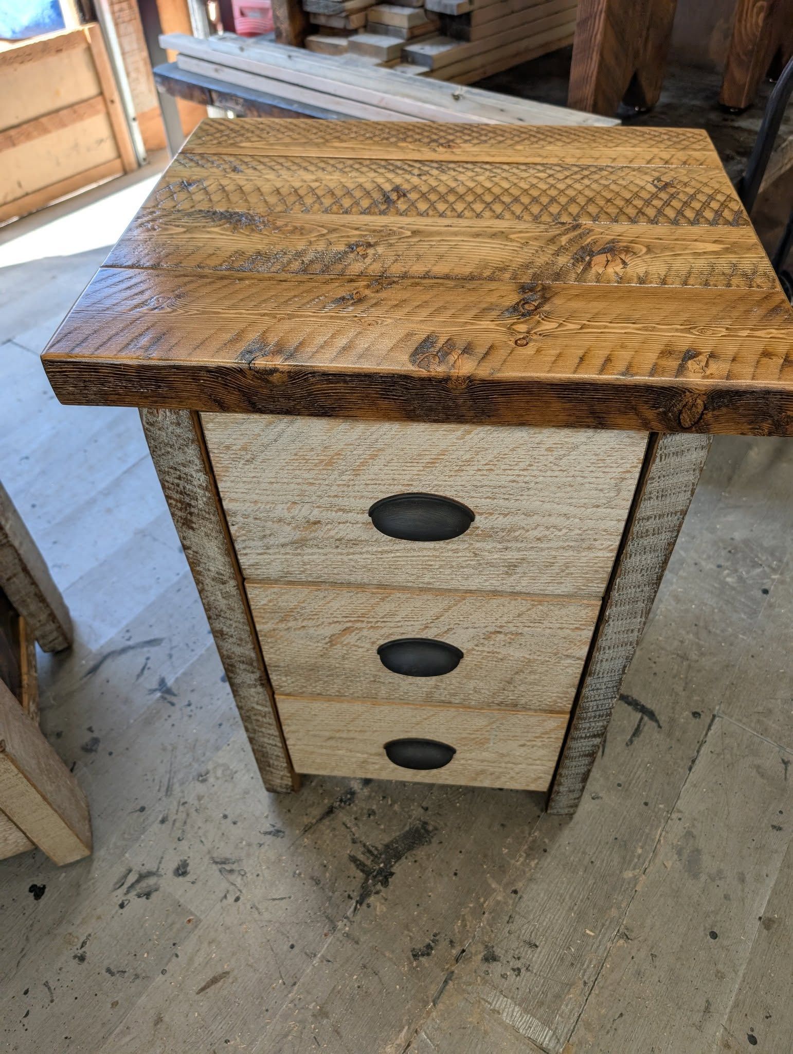 Rustic wooden cabinet with three drawers and a patterned top, standing on a concrete floor in a workshop.