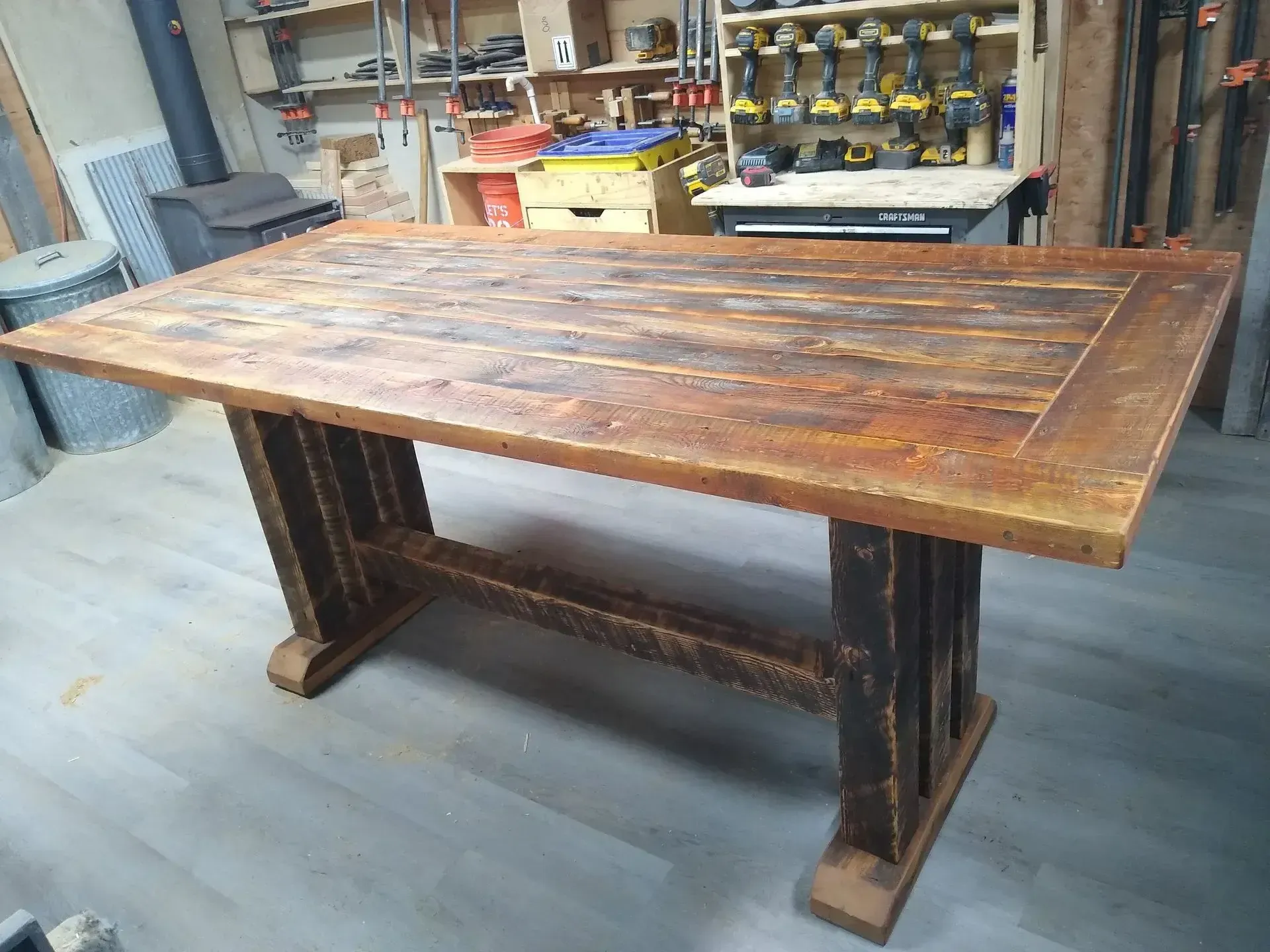 Rustic wooden dining table with dark pedestal legs in a workshop or garage.