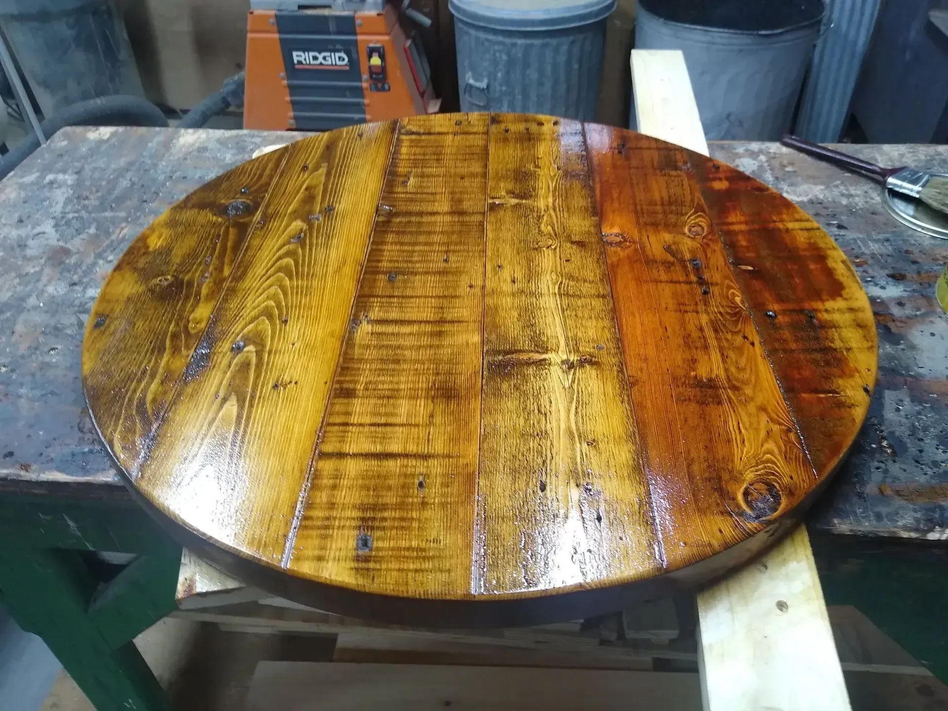 Round varnished wooden tabletop on a workbench, with glossy brown grain and wood shop tools in the background
