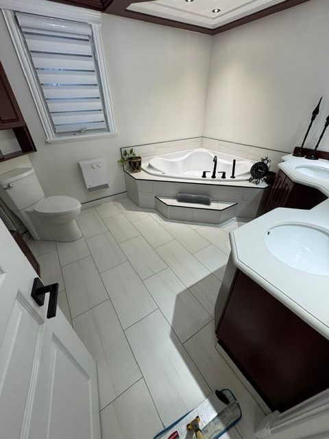 Salle de bain rénovée avec baignoire d'angle, toilettes, lavabo et fenêtre avec stores.
