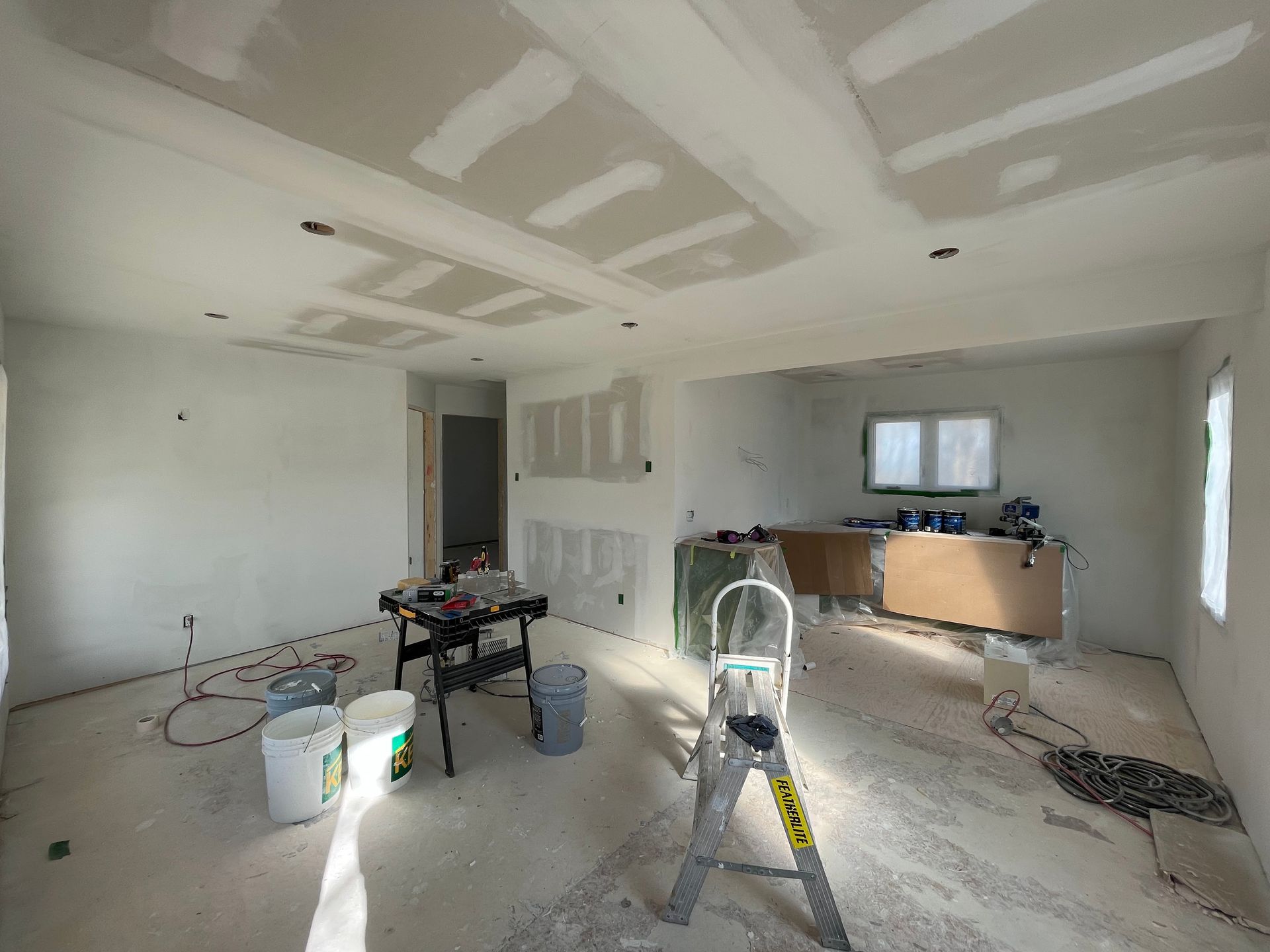 Interior of a room under construction with drywall, exposed beam ceiling, ladder, buckets, tools and construction materials.