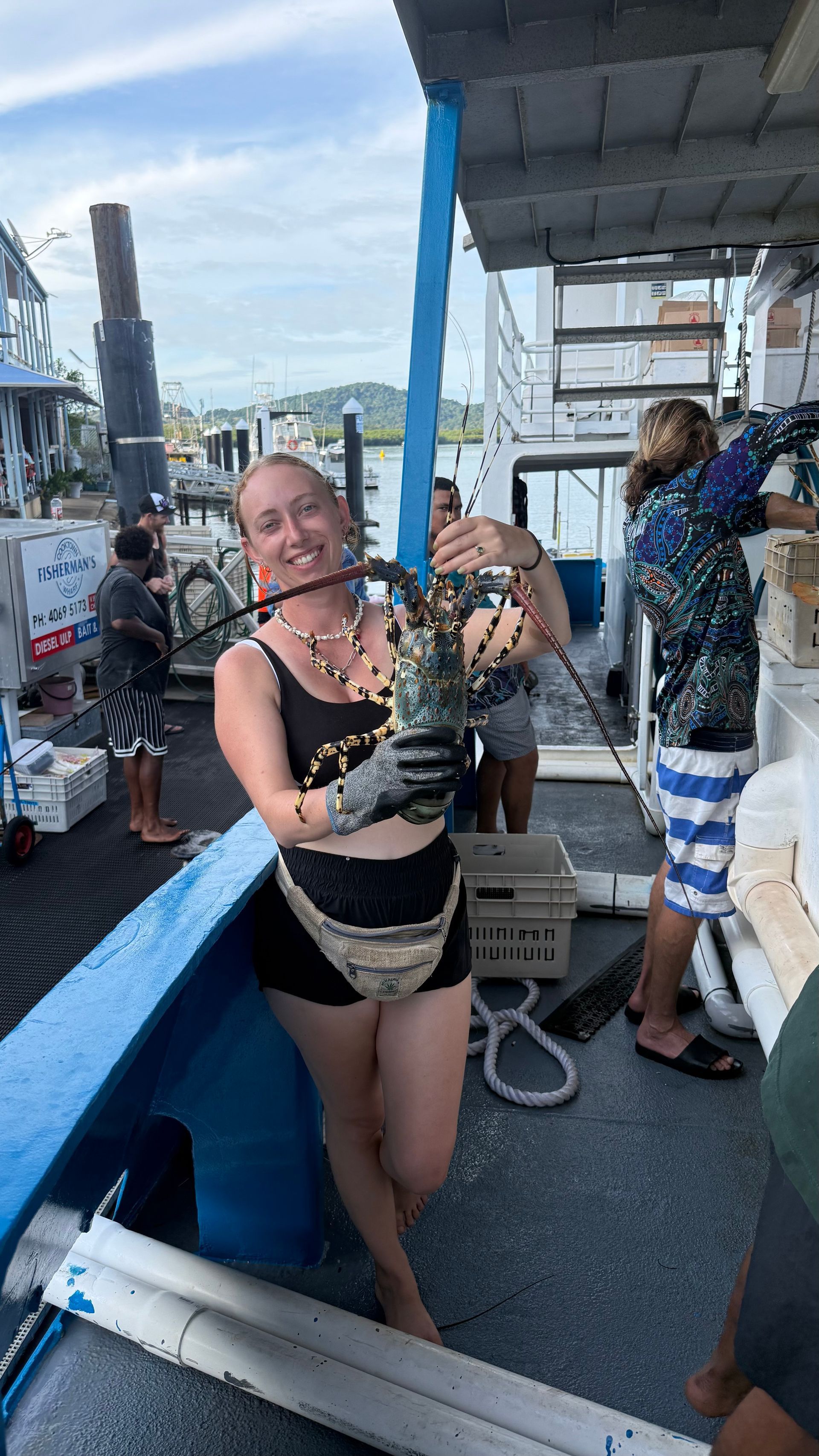 Jacinta holds a large crayfish on a blue commercial fishing boat docked at Cooktown, with crew in the background.