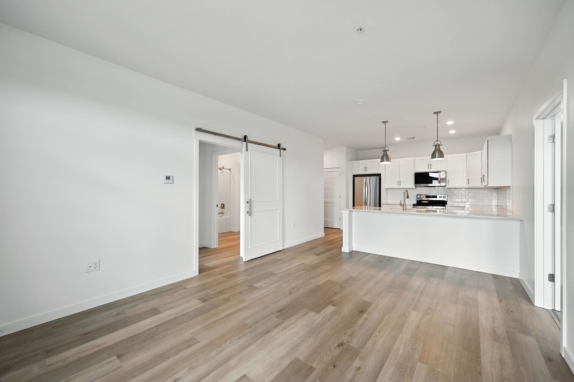 Open-concept apartment with hardwood floors, white kitchen, sliding barn door, and a door opening to a hallway.