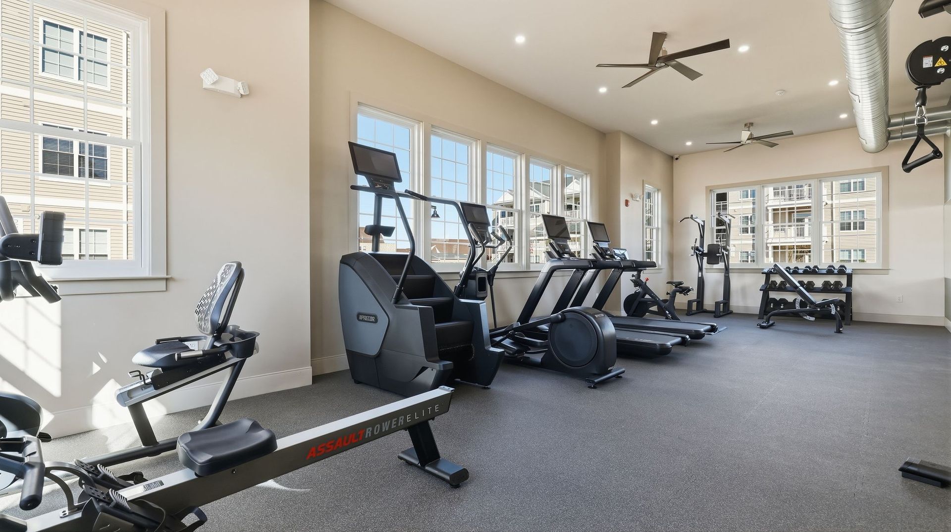Gym with exercise equipment, including treadmills, ellipticals, rowing machine, and free weights, in a room with windows.