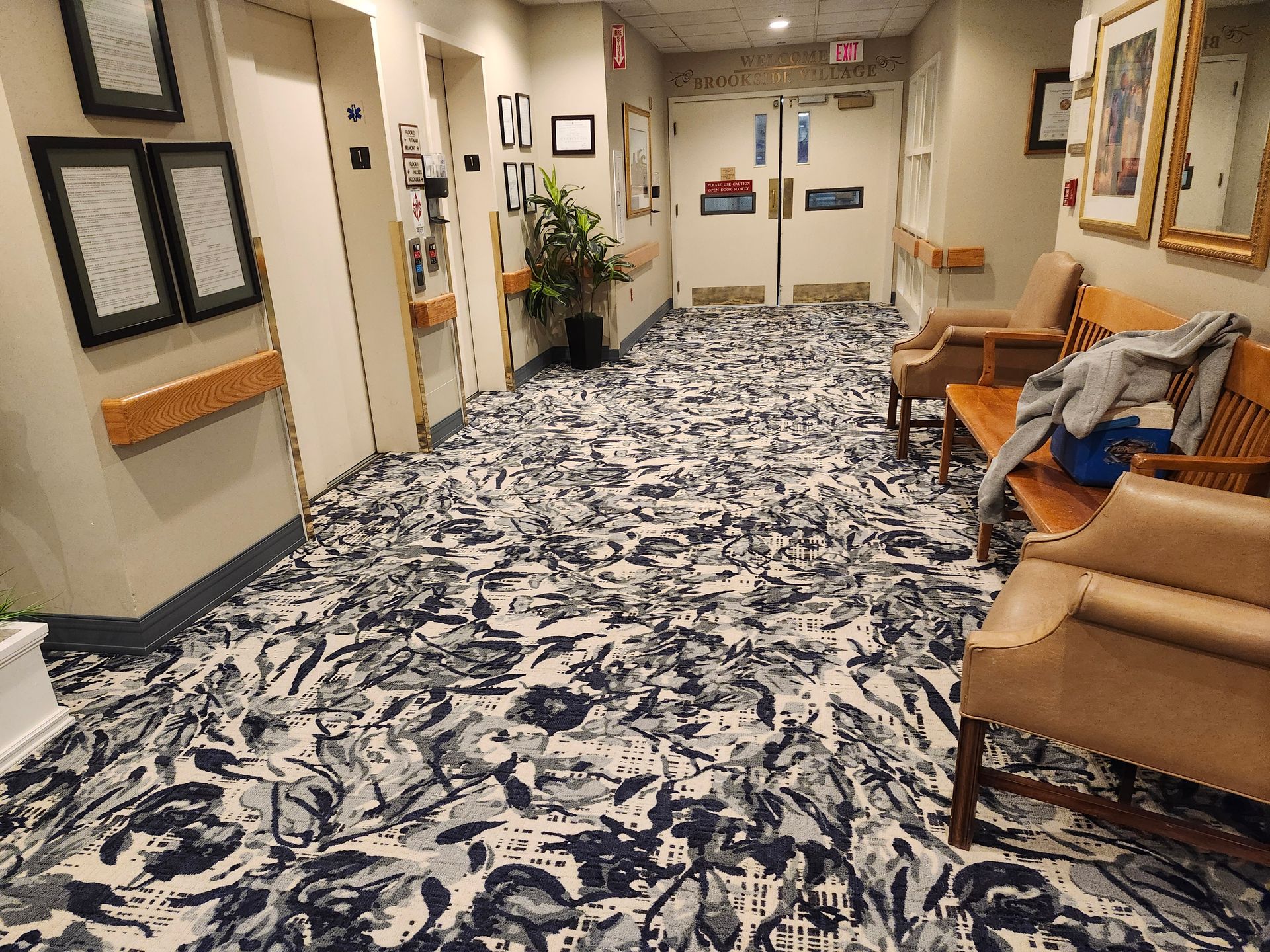 Empty hospital hallway with patterned carpet, wooden chairs, framed art, and double doors at the end.