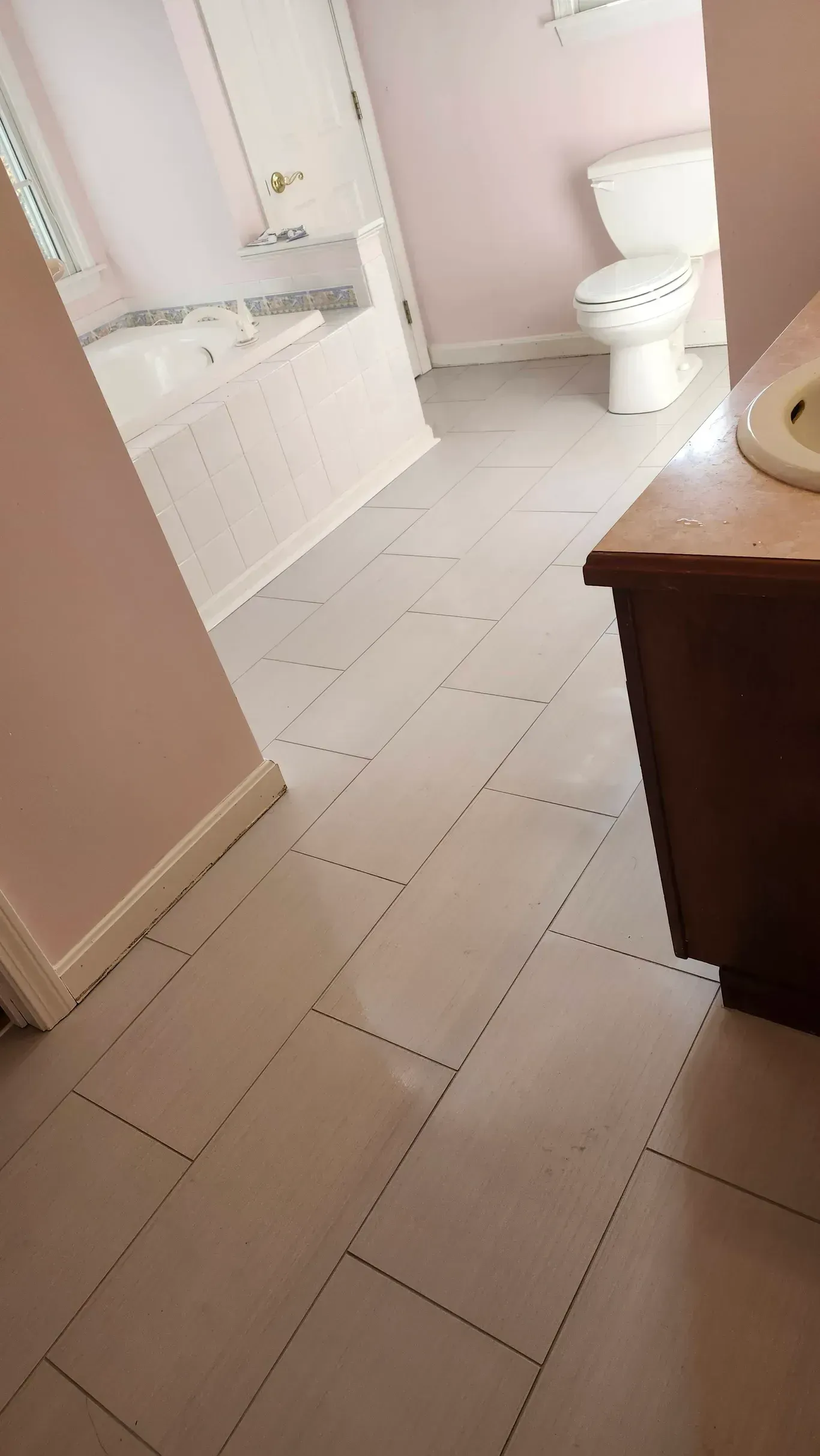 A bathroom with large, light-colored rectangular floor tiles, a white toilet, and a bathtub in the background.