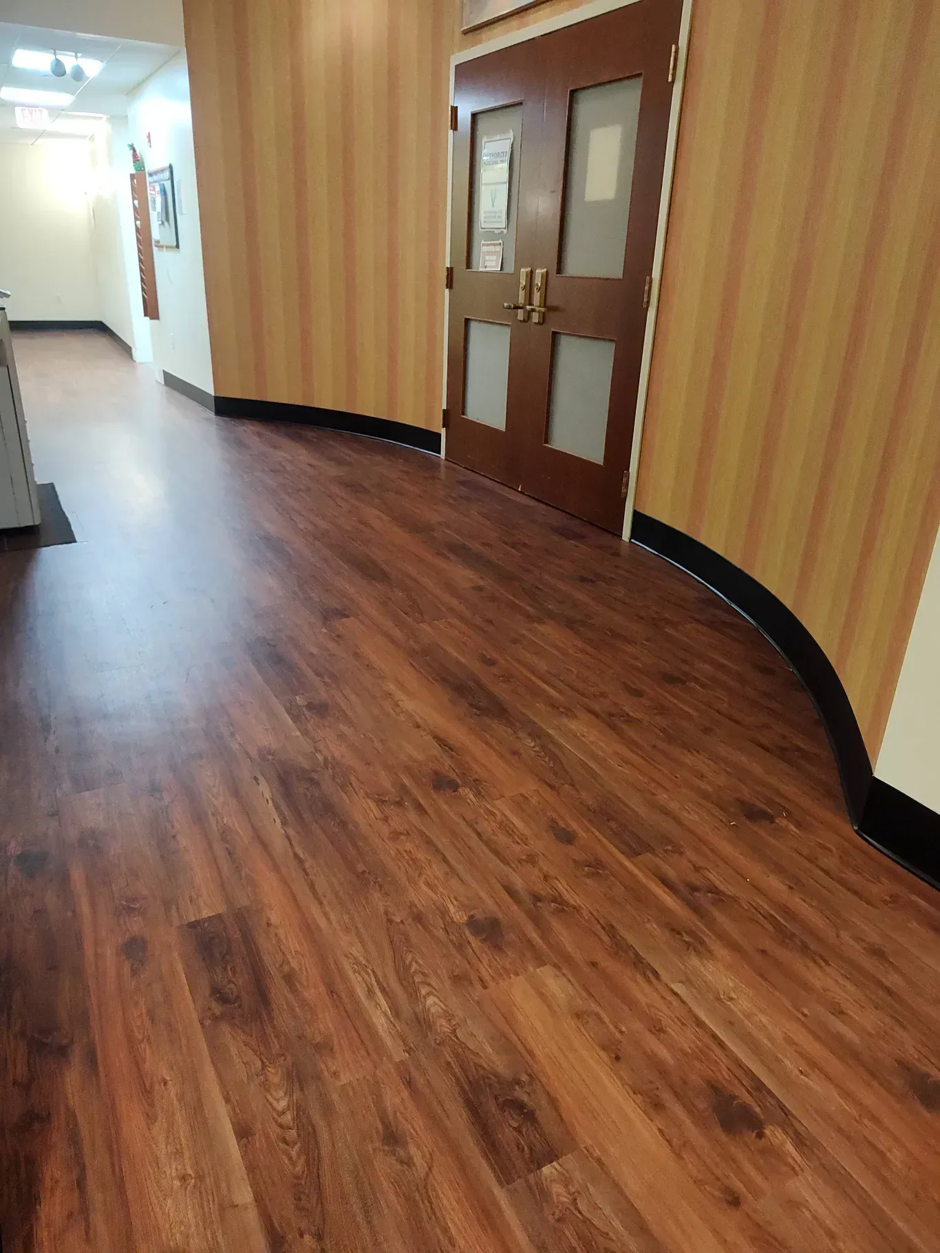 A hallway with wood-look flooring and striped wallpaper leads to a set of double wooden doors with glass panels.