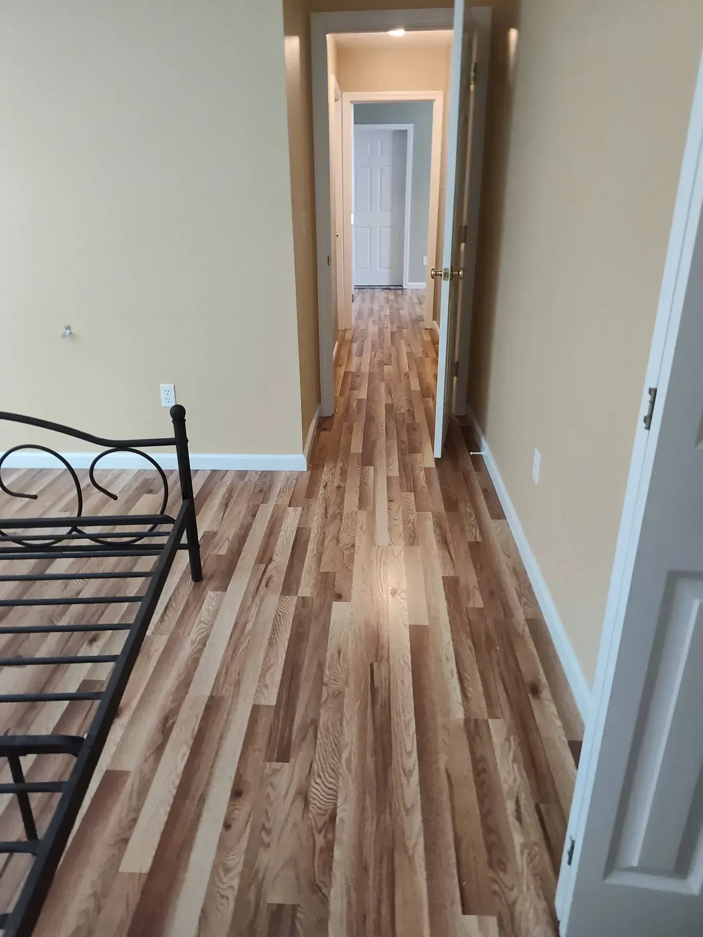 A room with light wood-plank flooring and tan walls, looking through an open doorway into a matching hallway.