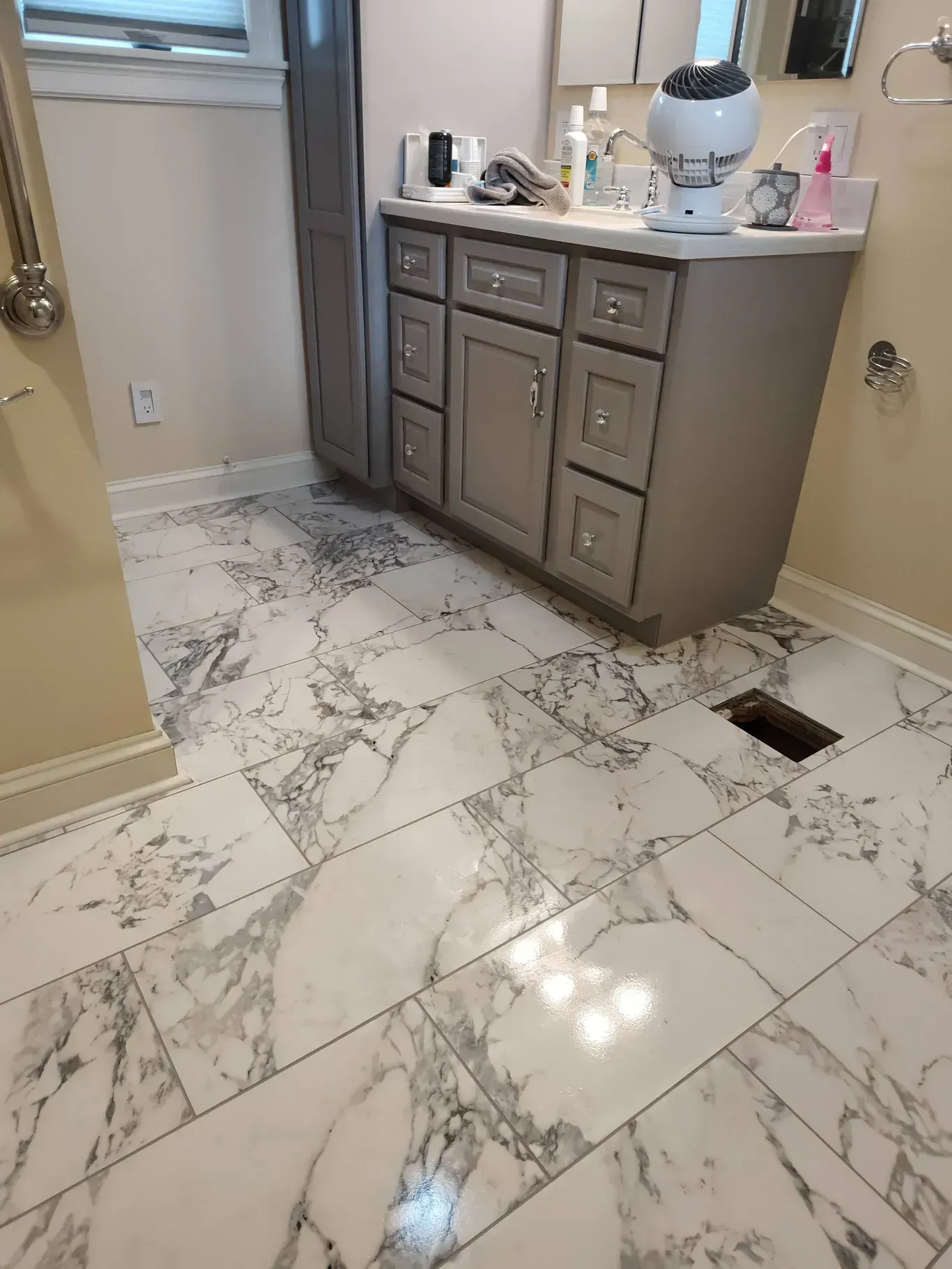 A bathroom vanity with a white countertop and grey cabinets stands on a floor featuring large white and grey marble tiles.