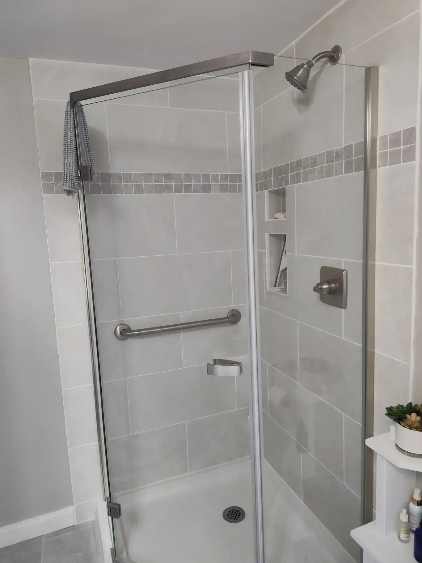 A walk-in shower with a glass door, light grey tiled walls, a metal grab bar, built-in shelving, and a chrome showerhead.