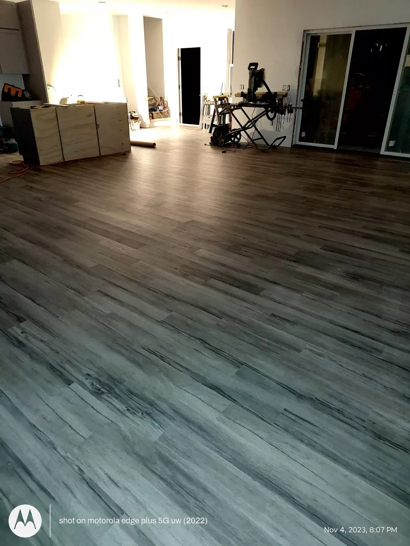 A room with grey wood-look flooring, showing an unfinished kitchen island and a miter saw on a stand in the background.