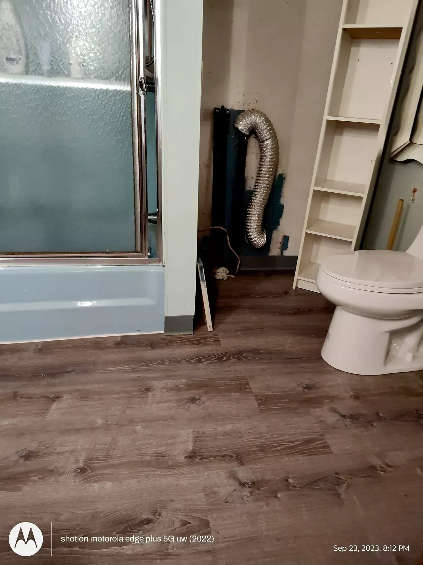A bathroom showing a shower, a toilet, a tall shelf, and a silver ventilation duct exposed on the wall.