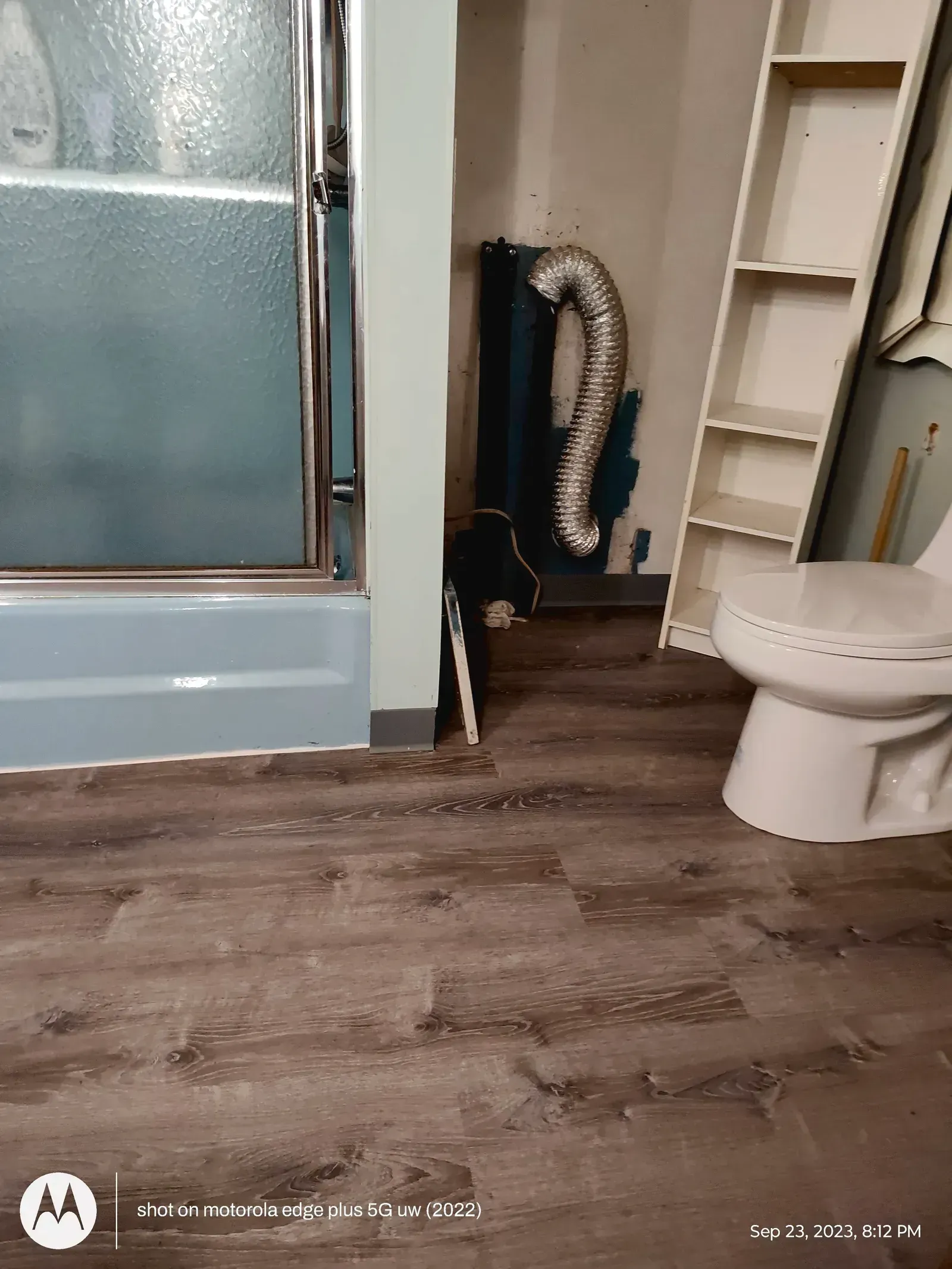 A bathroom featuring a shower stall, a toilet, a tall shelf, and a flexible metallic vent hose lying on the floor.