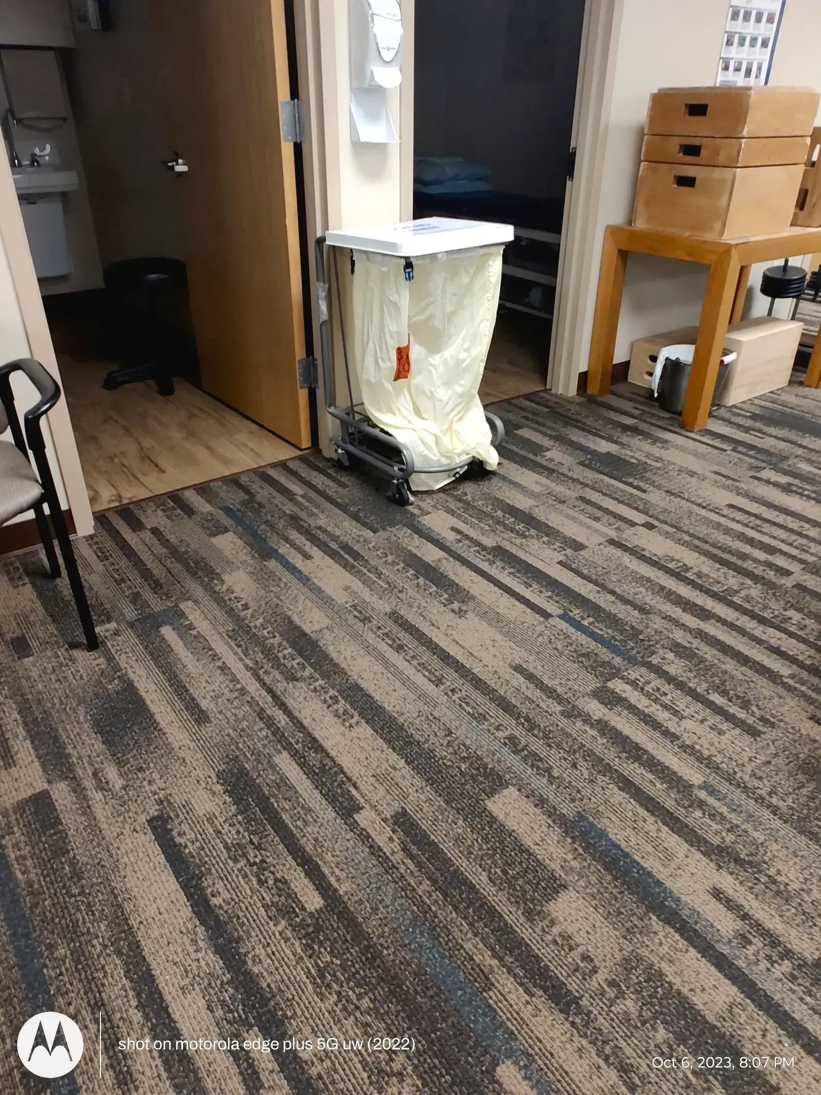 A white laundry cart stands on patterned carpet in a hallway near an open door and a wooden table with stacked boxes.