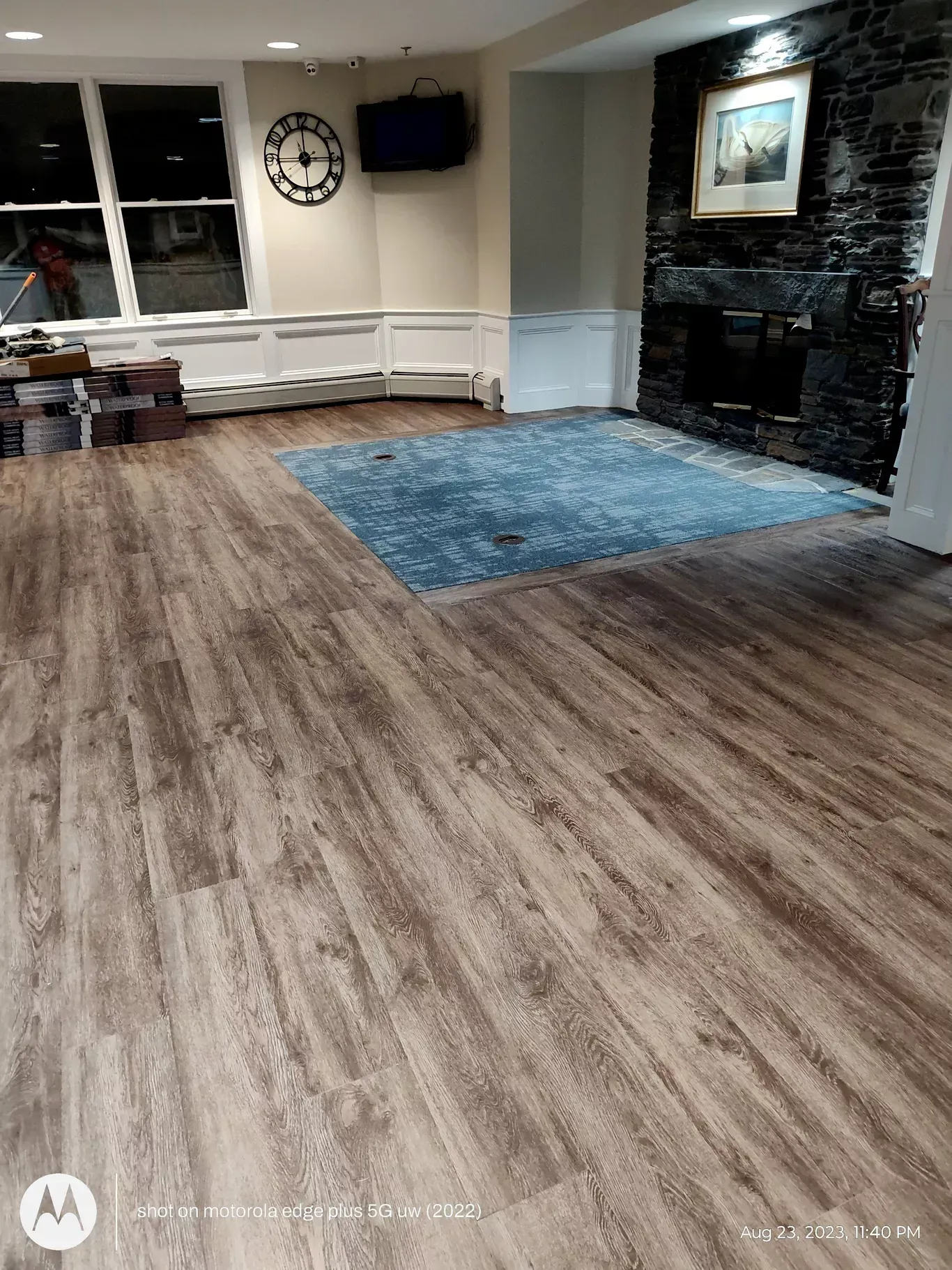 A room with wood-look flooring, a blue area rug in front of a stone fireplace, a wall clock, and white wainscoting.