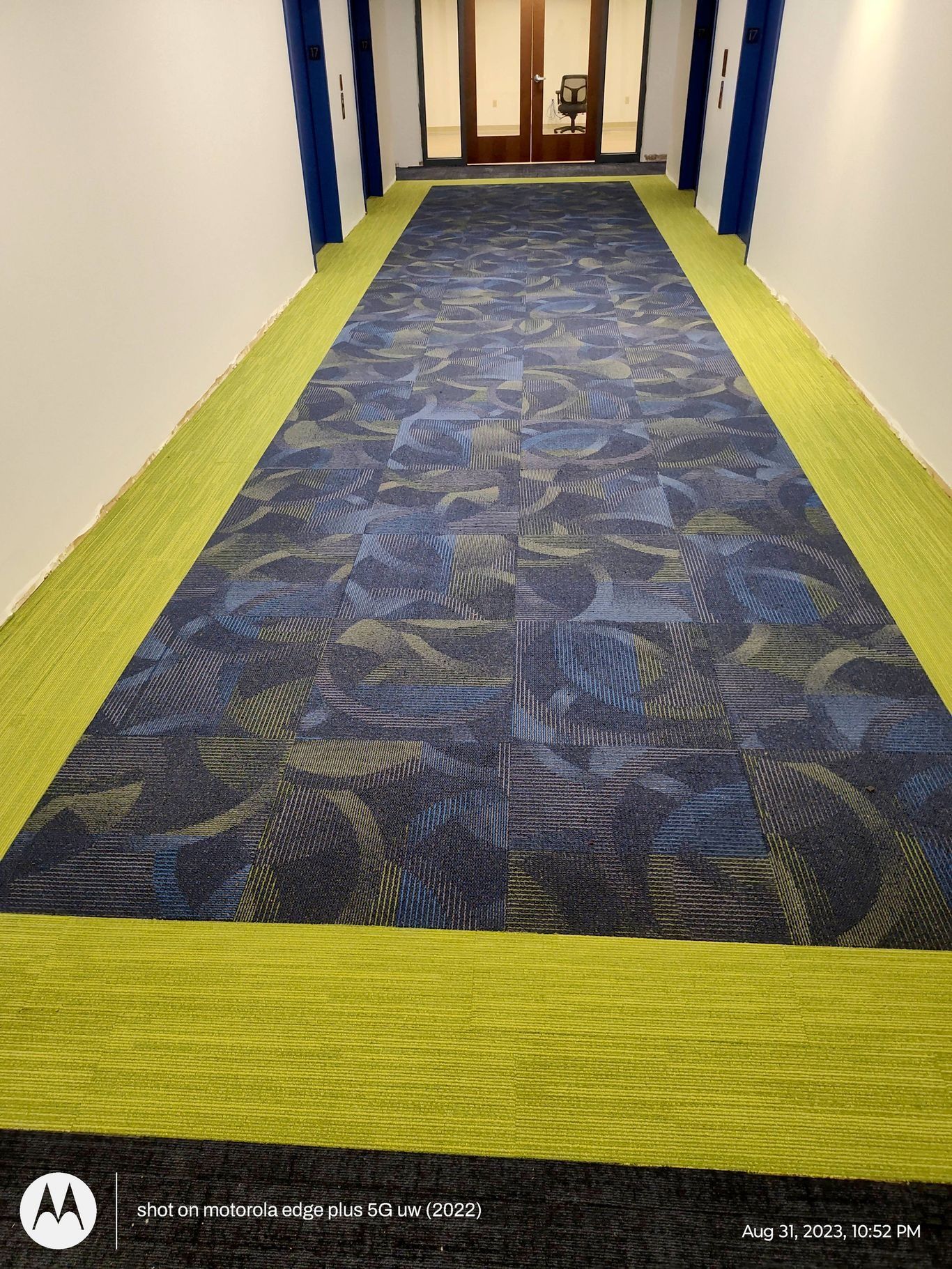 A long, narrow indoor hallway with a patterned blue rug bordered by bright yellow-green carpet leading to a glass door.