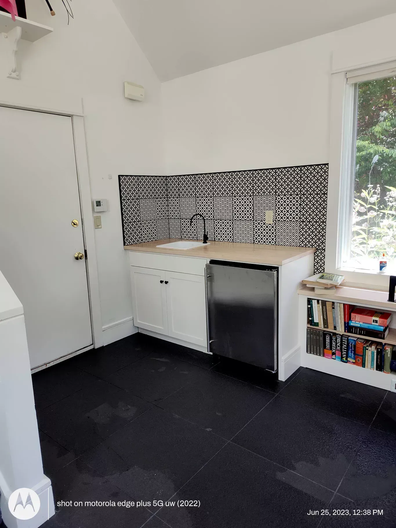 A kitchen area with white cabinets, a stainless steel dishwasher, black patterned backsplash, and black tiled floors.