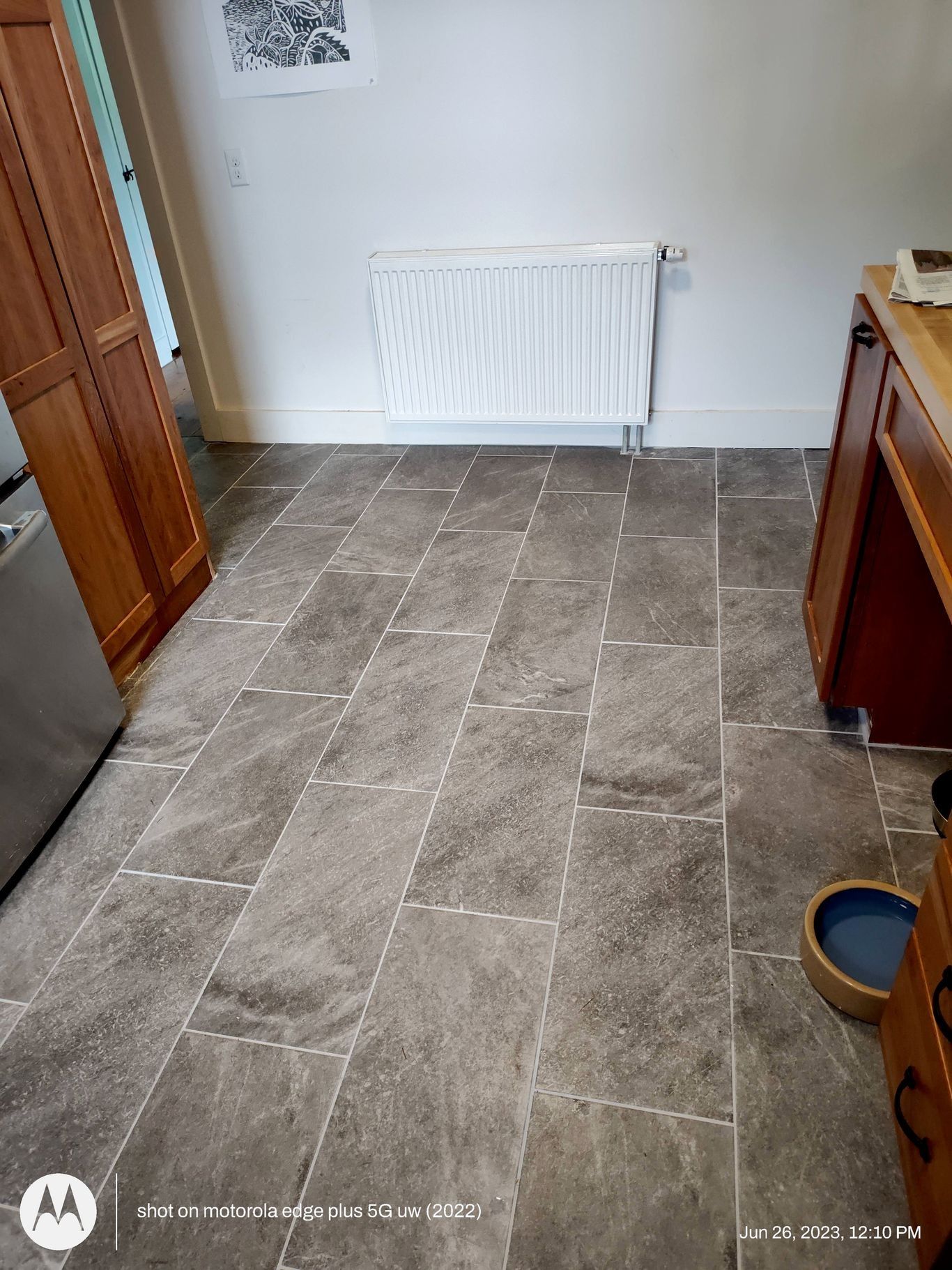 A kitchen floor with grey rectangular stone-look tiles, a white radiator on the back wall, and wooden cabinets on the side.